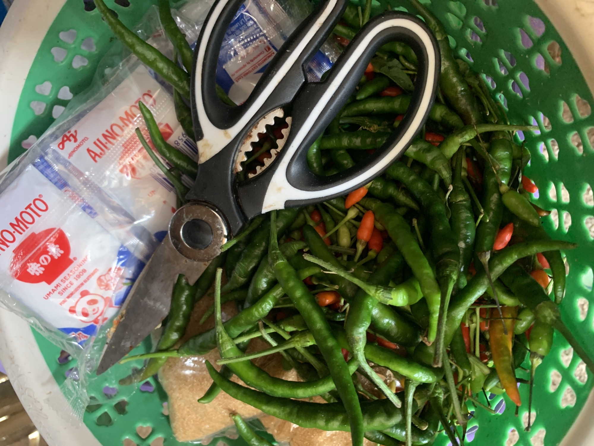 A high-angle, close-up shot captures a green plastic basket filled with numerous green and red chili peppers. A pair of black and white handled scissors lies on top of the peppers, with its blades open. Behind the scissors are two small, clear plastic packets of Ajinomoto seasoning, one with red lettering and a red circular logo featuring Japanese characters and a panda illustration, and the other with red and blue lettering. A bag of brown sugar is also visible to the left of the peppers. The basket itself is green and features a decorative pattern of heart-shaped cutouts. The scene is set in Kiblawan, Philippines.