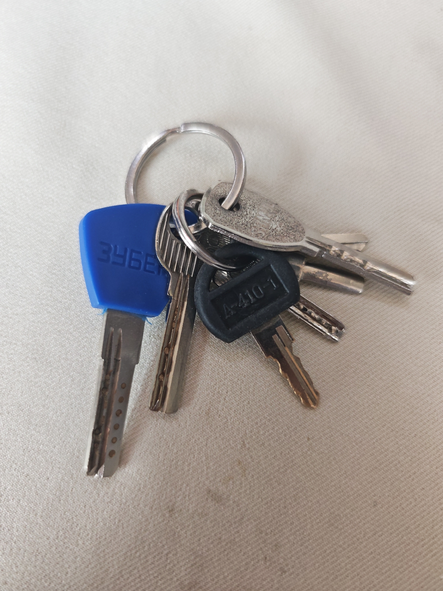 A keychain with several keys lies on a textured, off-white fabric surface. One key has a blue plastic head embossed with the Cyrillic letters "ЗУБЕ". Another key has a black plastic head with the marking "A-410-1". The keys are attached to a silver-colored metal keyring. The fabric surface is illuminated from above, casting subtle shadows from the keys. The location is unknown.