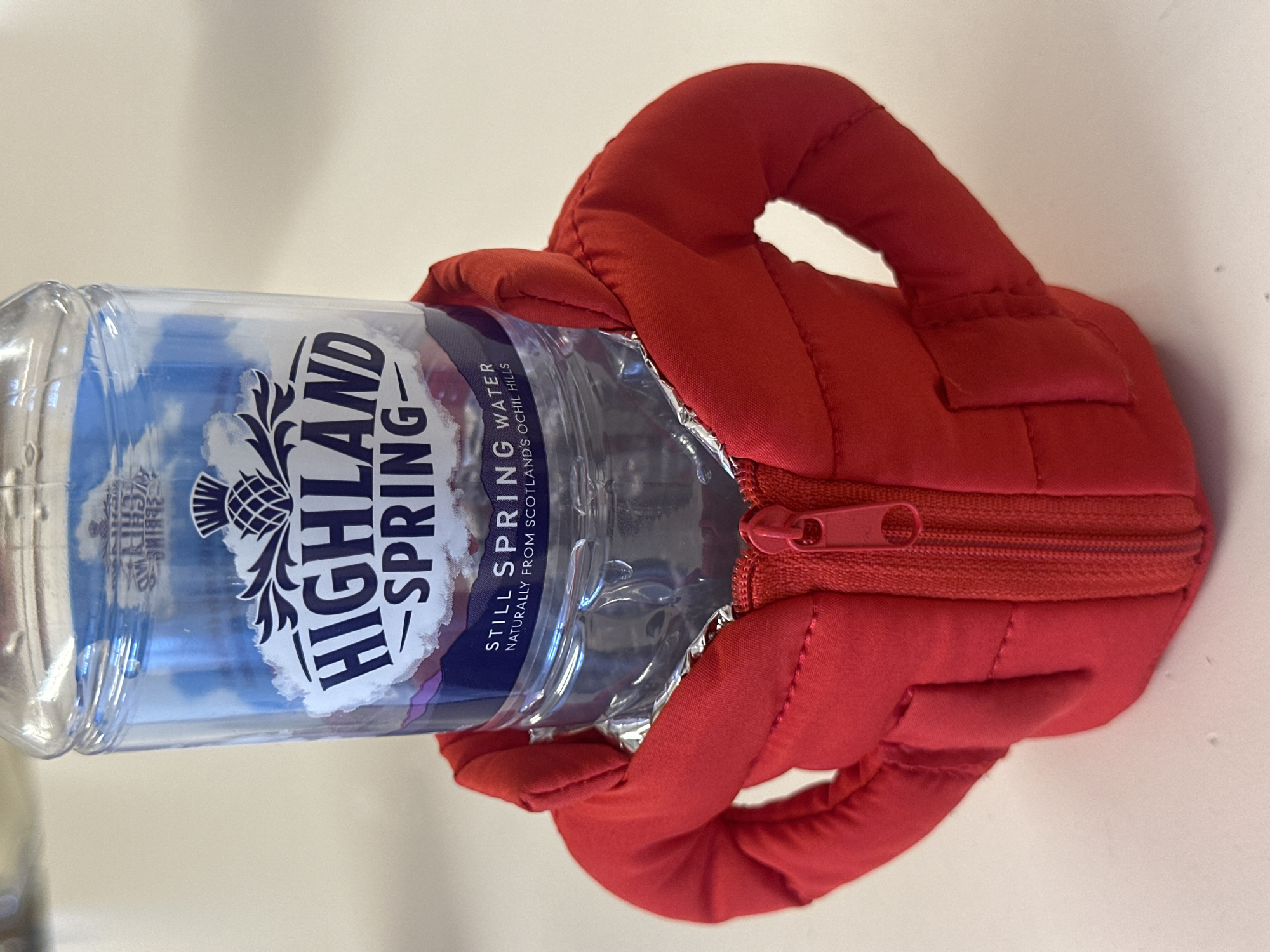 A clear plastic bottle of Highland Spring still spring water is depicted, encased in a vibrant red padded holder. The holder is designed to resemble a miniature puffer jacket, complete with a functional red zipper running down one side and a small, padded handle-like strap at the top.
The bottle's label is prominently visible, displaying "HIGHLAND SPRING" in large, dark blue lettering, along with a thistle emblem. Beneath the brand name, the text reads "STILL SPRING WATER" and "NATURALLY FROM SCOTLAND'S OCHIL HILLS," indicating its origin. The bottle appears full.
The objects are placed on a plain, light off-white or beige surface, suggesting an indoor setting such as a table or counter. The lighting is uniform, implying an indoor environment, possibly during the daytime. No individuals are present in the image, and no specific activity is occurring beyond the static presentation of the water bottle within its holder. The image itself is oriented horizontally, with the bottle's base to the right.