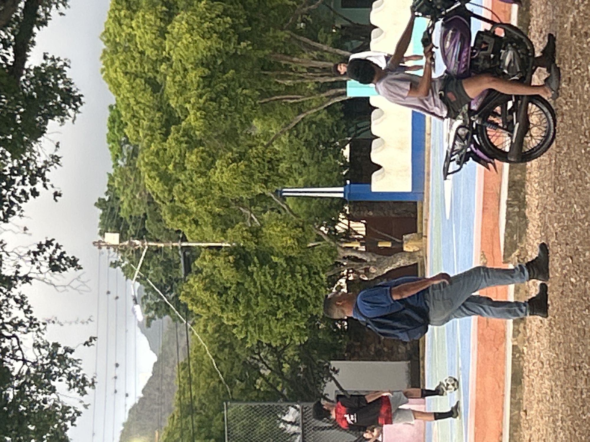 The image depicts an outdoor scene in Santa Ana, Venezuela, possibly a park or a sports complex. In the foreground, a young man wearing a purple t-shirt and dark shorts sits on a purple motorcycle. To his left, a man in a blue shirt and jeans walks with a backpack. Further in the background, another young man in a black and red shirt and grey shorts stands near a soccer ball on a blue and red court. The environment features lush green trees, mountains in the distance under a cloudy sky, and a fence enclosing a sports area.