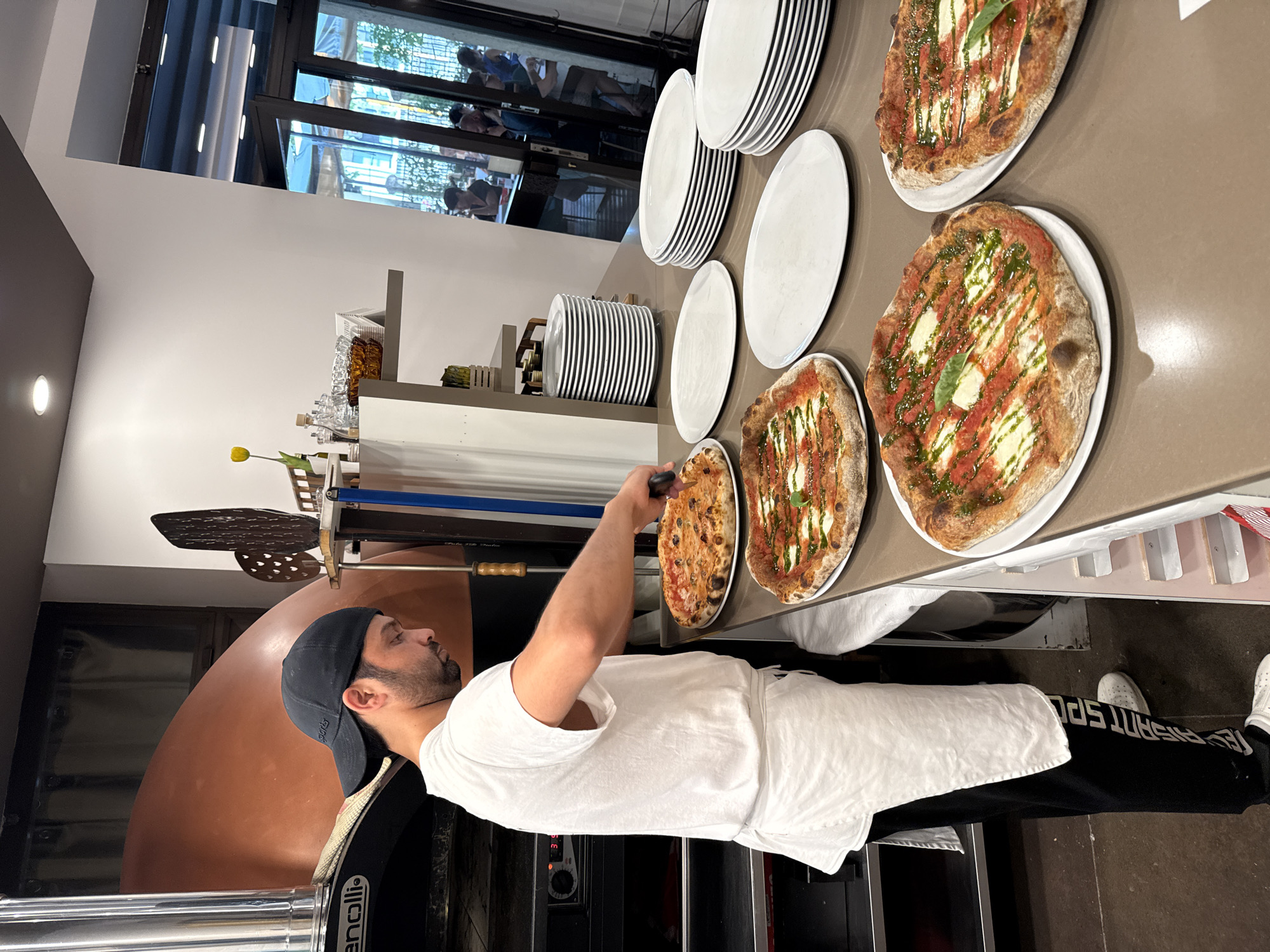 A male pizza maker, wearing a black baseball cap, a white t-shirt, and a white apron over black sweatpants with white lettering, is standing behind a counter in what appears to be a pizzeria. He is in the process of cutting a pizza with a pizza cutter. In front of him, on the counter, are several finished pizzas on white plates, and stacks of white plates. Behind him, to the left, is a large, dome-shaped pizza oven. In the background, through a large glass door, patrons are seated at tables in an outdoor dining area. The scene is consistent with a pizzeria in Trento, Italy, a city known for its culinary scene and numerous pizzerias.