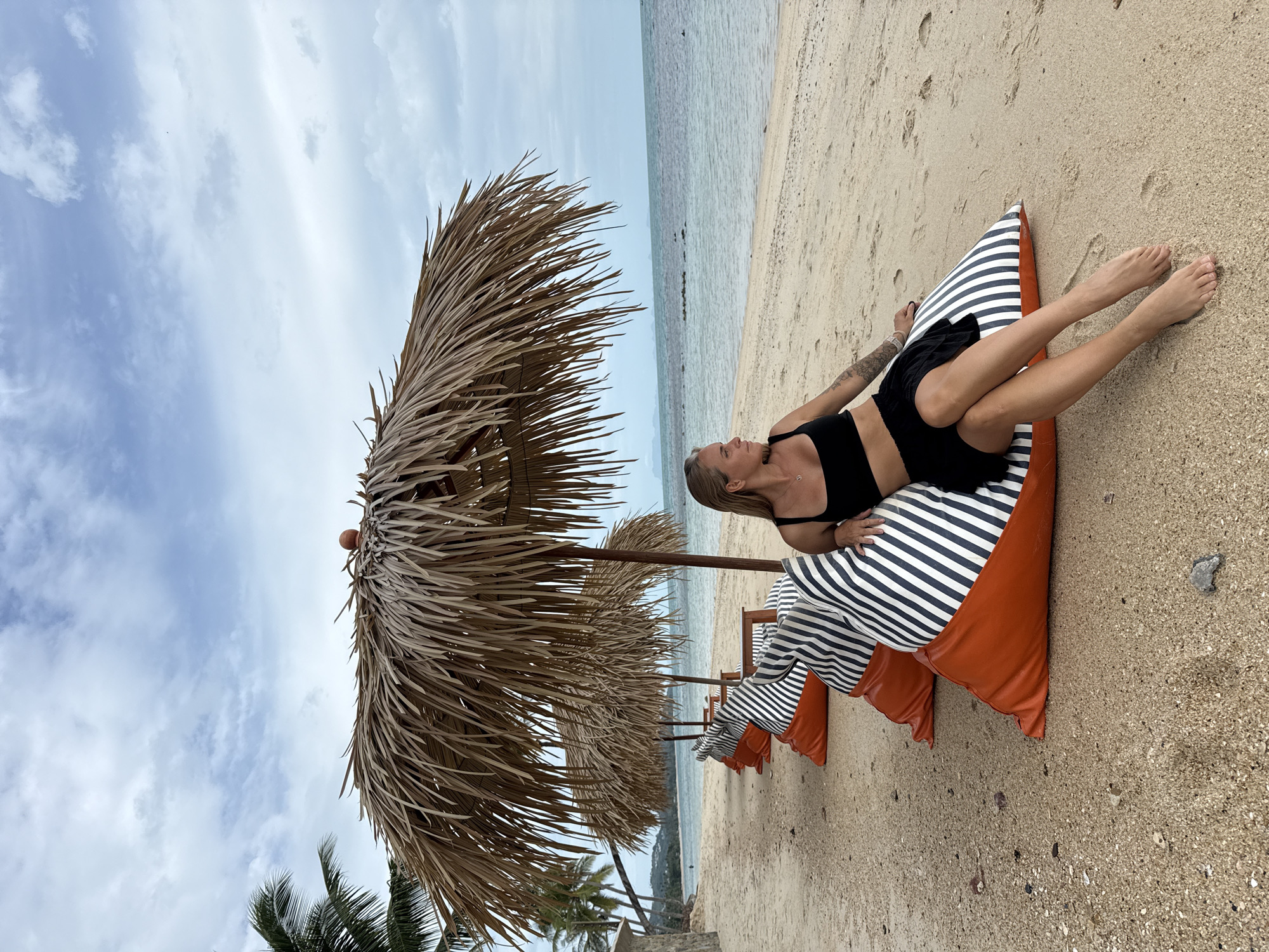 A blonde woman with tattoos on her arm sits on a large, orange and striped beanbag chair on a sandy beach in Ko Samui District, Thailand. She wears a black bikini top and a black skirt. Behind her are three more beanbag chairs and thatched umbrellas providing shade. The ocean is visible in the background, with a cloudy sky overhead. Palm trees are visible to the left of the frame. The sand is littered with small pebbles and shells.