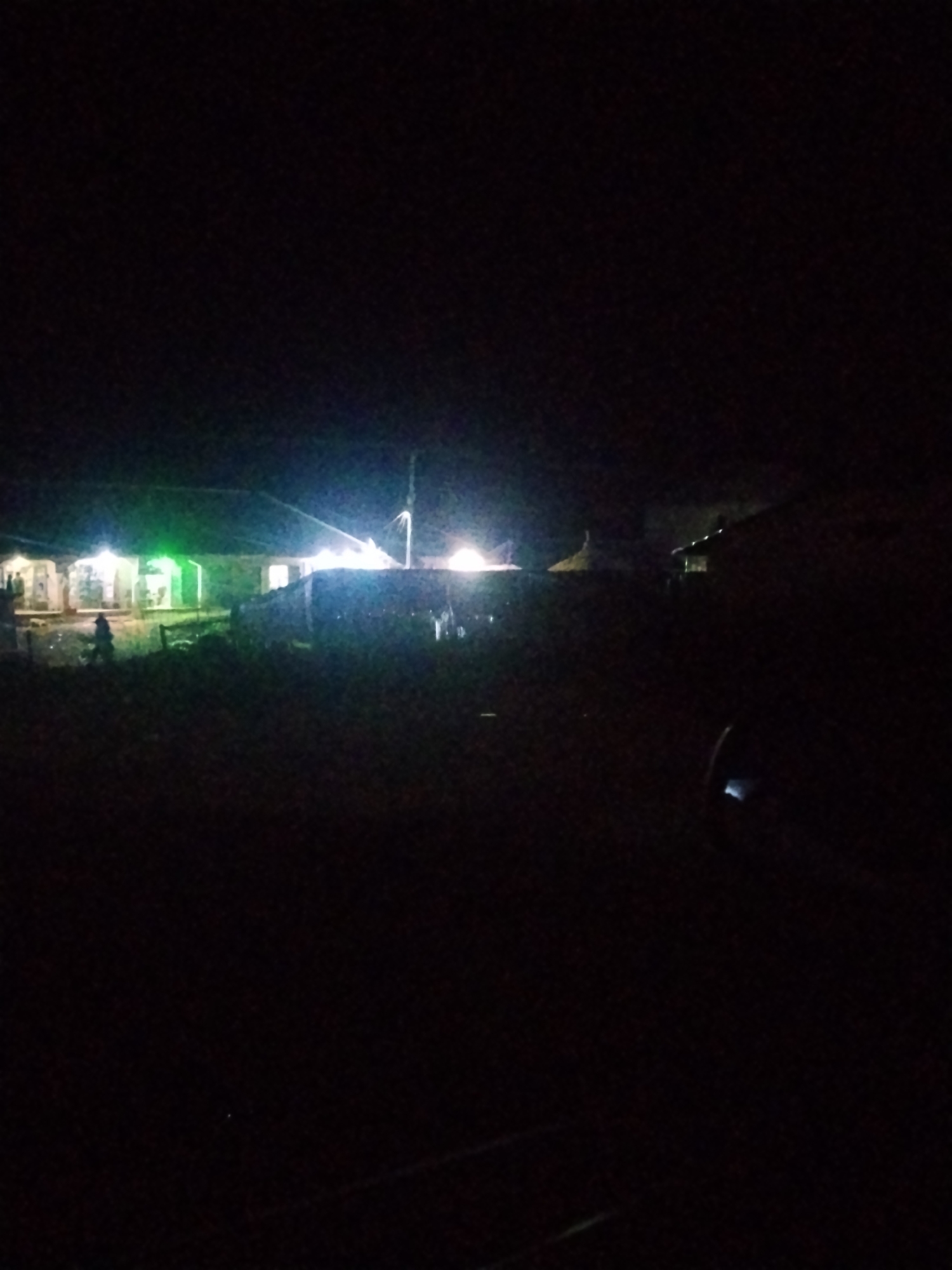 The image depicts a dimly lit outdoor scene at night, possibly in the city of Durumi, Nigeria. Several structures with illuminated windows are visible, suggesting human habitation or activity. The lighting is primarily from artificial sources, casting a faint glow on the surrounding environment. There are indications of vegetation or rough terrain in the foreground, with shadows obscuring finer details. The overall impression is one of a quiet, sparsely populated area at night, with some buildings lit up.