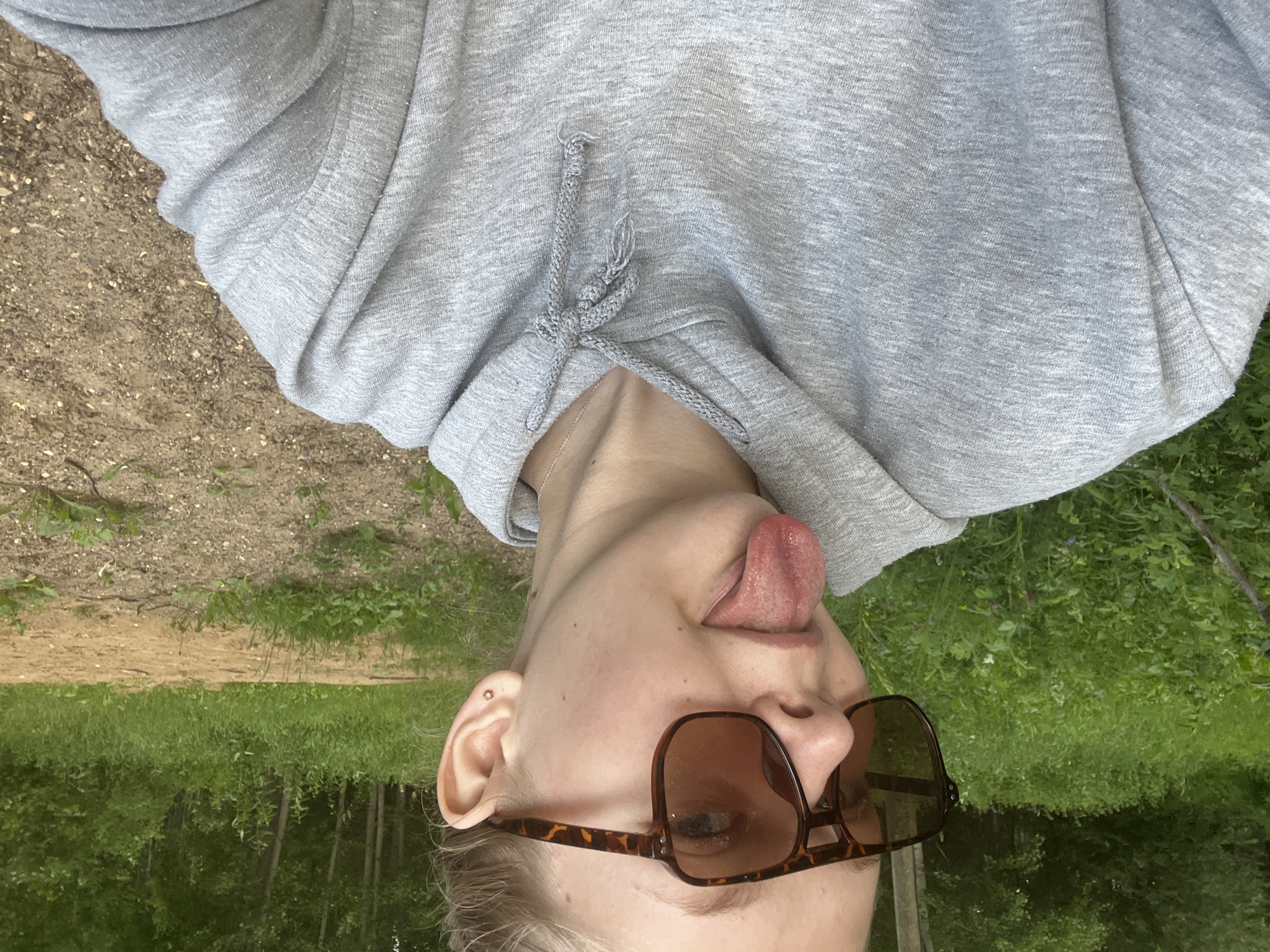 This is a close-up, upside-down selfie of a person with fair skin. The person is wearing a gray hooded sweatshirt with drawstrings and a silver necklace. They are also wearing brown, tortoise-shell patterned sunglasses. The person's tongue is sticking out.
The background appears to be an outdoor scene with greenery, suggesting a natural setting like a park or forest. There's also a sandy path visible. The lighting suggests it might be daytime, and the greenery implies a temperate season. The overall mood conveyed by the image is playful and candid. No text is visible in the image. The provided location context is Klepiki, Russia.
