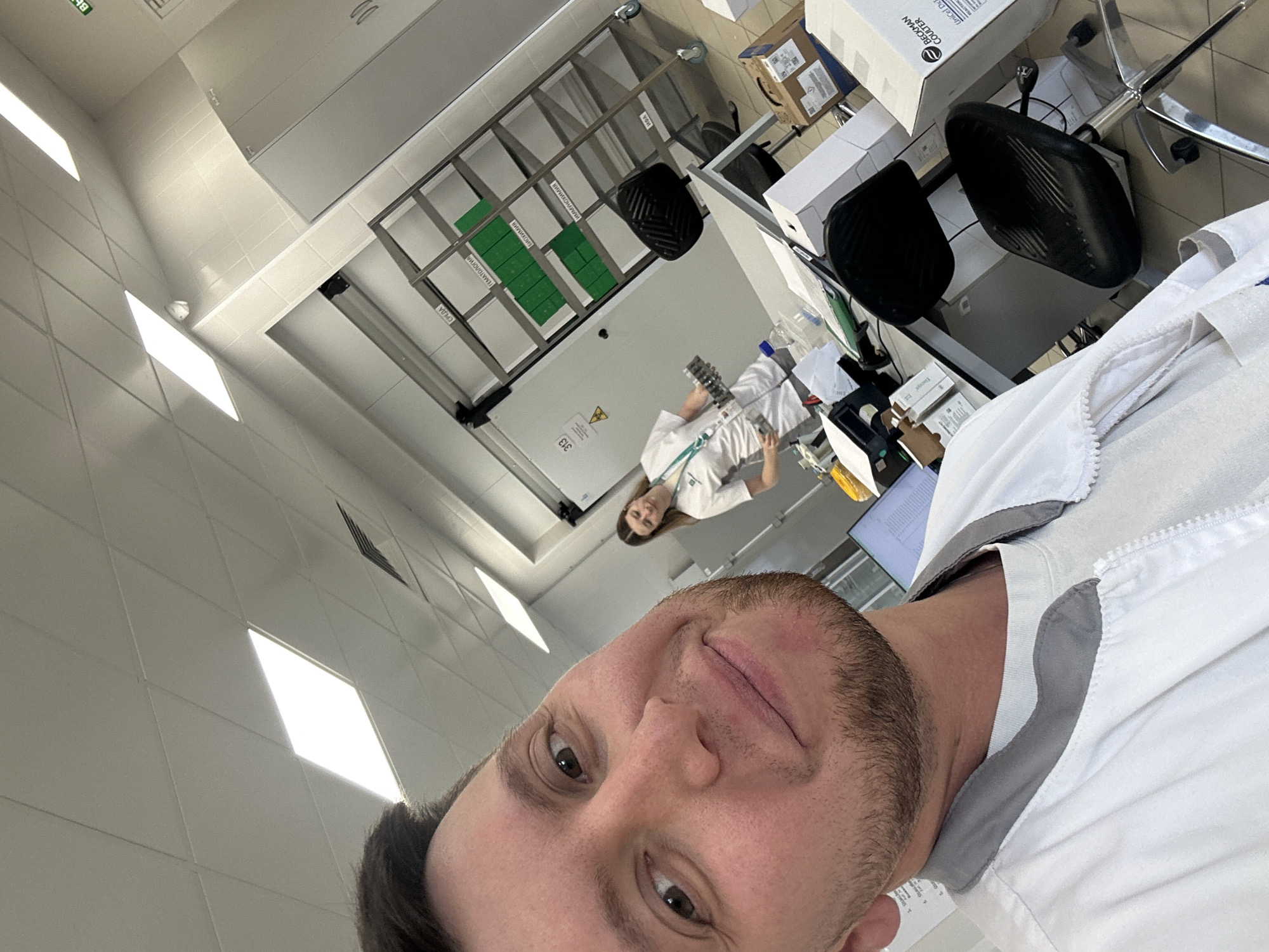 A selfie photograph shows a man with light brown hair and a short beard wearing a white lab coat in a lab setting. In the background, another person, a woman, is also in a lab coat and is standing near a desk with lab equipment. Behind her is a white wall with labeled racks. Boxes and chairs are visible. The setting appears to be a scientific or medical laboratory. The location is unknown; there is no evidence to suggest Moscow, Russia.