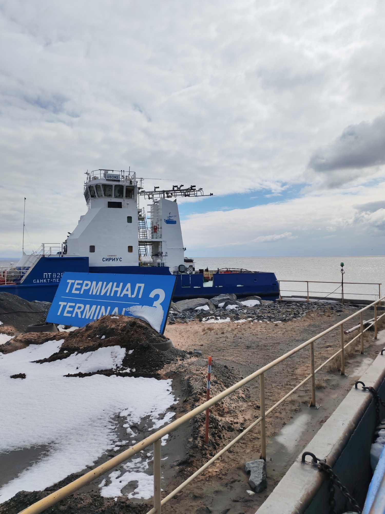 An industrial waterside scene in Shcheleiki, Russia, depicts a large blue and white vessel, likely a tugboat or icebreaker, moored or beached near the shore under an overcast sky. The vessel features a prominent white superstructure with multiple decks and windows. Text visible on the vessel includes "UBOM3" on the top of the bridge, "СИРИУС" (Sirius) on its side, and "ПТ 829" and "САНКТ-ПЕТ" (likely Sankt-Peterburg or St. Petersburg) towards the bow, indicating its home port. A logo of a stylized boat is also present.
In the foreground, a blue sign with white text is partially covered by snow and dirt, displaying "ТЕРМИНАЛ" (Terminal) in Cyrillic, "TERMINAL" in English, and the large number "3". The ground is a mix of dirt, gravel, and melting snow, suggesting late winter or early spring conditions. A light-colored metal railing runs across the lower right of the frame, bordering a concrete edge with chains. Beyond the shore, a wide body of water extends to the horizon under a sky filled with grey and white clouds. The scene is devoid of people and appears calm and functional.