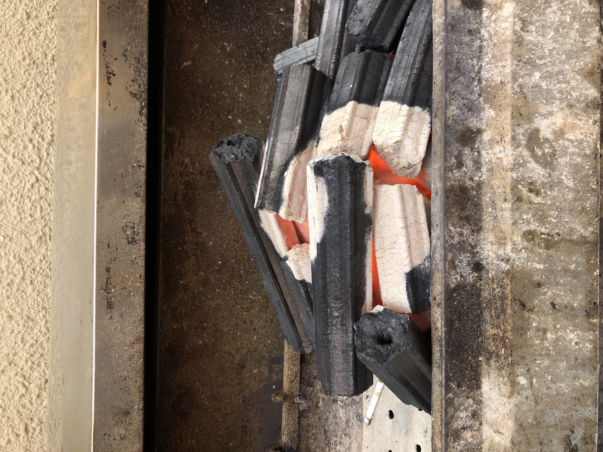 The image shows a pile of charcoal briquettes, some of which are glowing red with heat, indicating they are burning. The briquettes are dark gray to black on the outside and appear to have a white or light gray coating on some surfaces. They are arranged in a haphazard pile within a metal grill or fire pit. The surrounding metal is weathered and shows signs of rust and soot. The background consists of a textured wall, possibly stucco, and a metallic surface above the grill. The scene is likely located outdoors, possibly in a residential or commercial area of Đà Nẵng, Vietnam, given the context. There are no people or animals visible in the image.