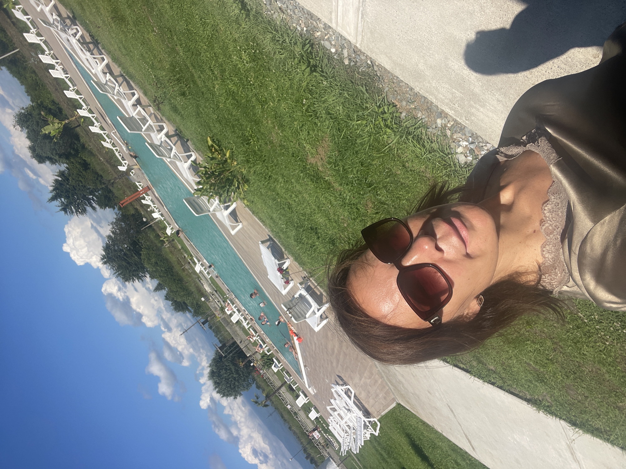 A woman in sunglasses and a satin top takes a selfie outdoors near a swimming pool. The pool is surrounded by lounge chairs and green grass. In the background, trees and a clear blue sky with white clouds are visible. Several people are swimming or relaxing in the pool. The setting appears to be a resort or hotel in Adzyubzha, Abkhazia.