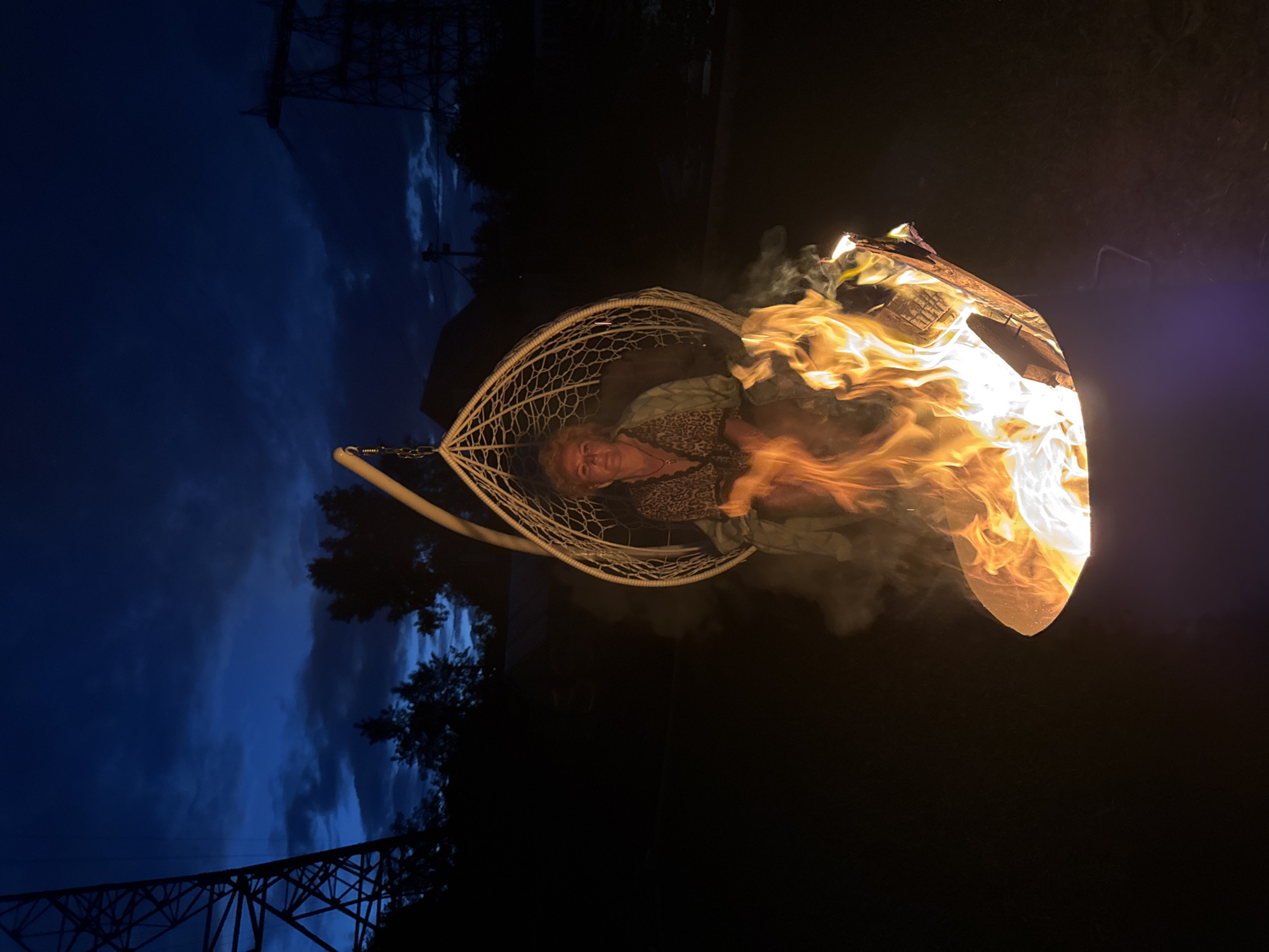 A woman sits in a hanging chair next to a fire pit at dusk. The sky is a deep blue with scattered clouds. In the background, there are dark trees and what appear to be power lines. The fire pit is in the foreground and is filled with bright flames. The woman is wearing a leopard print top and a light green robe. She is looking at the camera. The hanging chair is made of white woven material and is suspended from a metal frame. The fire pit is a dark metal basin. The overall impression is one of a cozy, relaxing evening outdoors.
