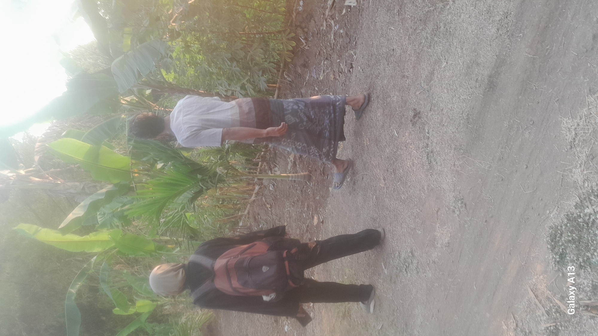 The image depicts two individuals walking on a dirt path, likely in the rural outskirts of Hadipolo, Indonesia. Lush green vegetation, including banana trees, dominates the background, with sunlight casting a soft glow.

The person on the left, facing away from the camera, wears a headscarf, a dark top, dark pants, and a backpack. They are holding a phone in their left hand. The person on the right, also facing away, is wearing a striped t-shirt and a patterned sarong. Both individuals are wearing sandals and appear to be walking in the same direction. The path is unpaved and appears dry and dusty. The overall atmosphere suggests a warm, possibly tropical, environment. The "Galaxy A13" watermark at the bottom indicates the image was captured with a Samsung Galaxy A13 smartphone.