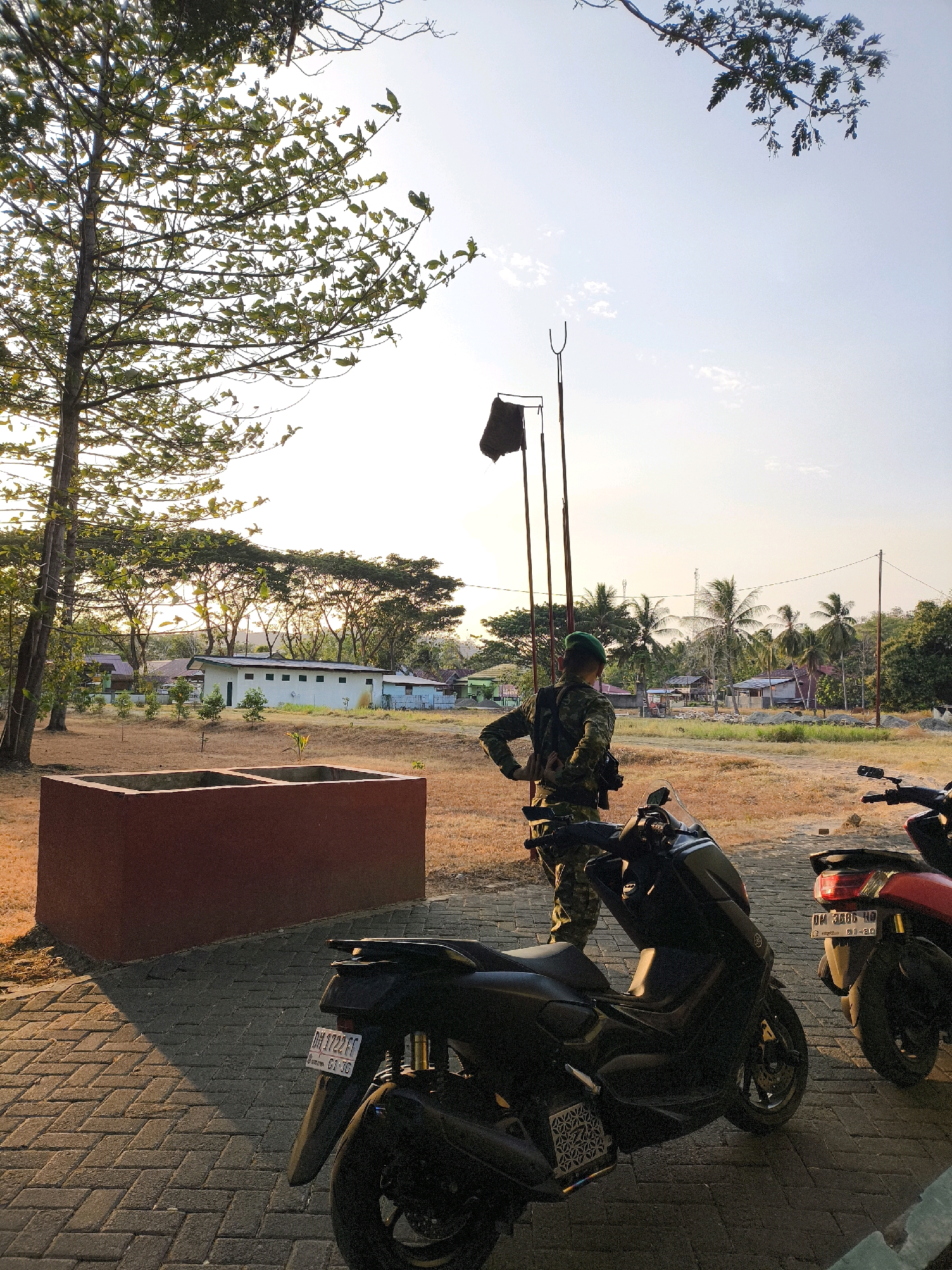 In a scene set in the city of Unknown, Indonesia, a man in camouflage military uniform stands near two parked scooters. The man faces away from the camera, with his left hand on his hip, his right hand adjusting something on his belt. He wears a green cap and a backpack.
The scooters are parked on a paved area made of interlocking bricks. The scooter in the foreground is black, with a license plate that reads "DM 1722 FF". The scooter to its right is red, with a license plate that reads "DM 3686 HD" and a sticker displaying "01-26".
Behind the scooters, a dry, grassy field is visible, with a rectangular concrete structure in the middle ground. In the background, a white building with multiple windows is situated amongst trees. Further in the distance, houses and palm trees are scattered across the landscape. The sky is clear, with the warm light of the setting sun casting long shadows. A tall tree with green leaves stands to the left of the frame. A flag is visible on a pole in the background.