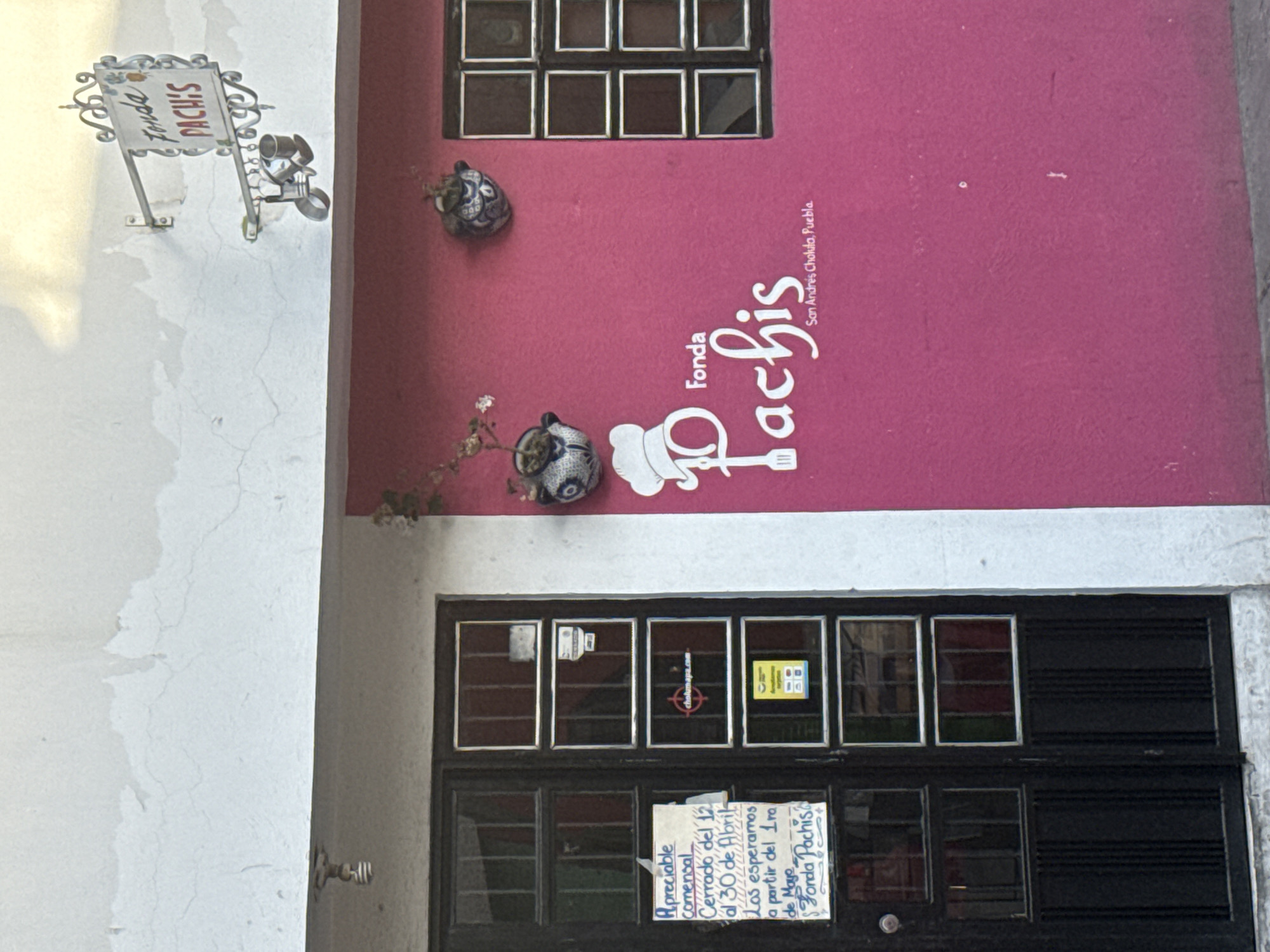 The image depicts the exterior of a restaurant named "Fonda Pachis" located in San Andrés Cholula, Puebla, Mexico. The building has a pink facade with a white logo and address. A black double door with multiple square glass windows makes up the entrance. A handwritten sign taped to the door states that the restaurant is closed from April 12th to April 30th, and will reopen on May 1st. There are two decorative ceramic pots mounted on the wall, one above and one to the side of the logo. A white wall adjoins the pink building, with a metal sign extending from the wall that also reads "Fonda Pachis." A small black-framed window is visible on the upper part of the pink wall.