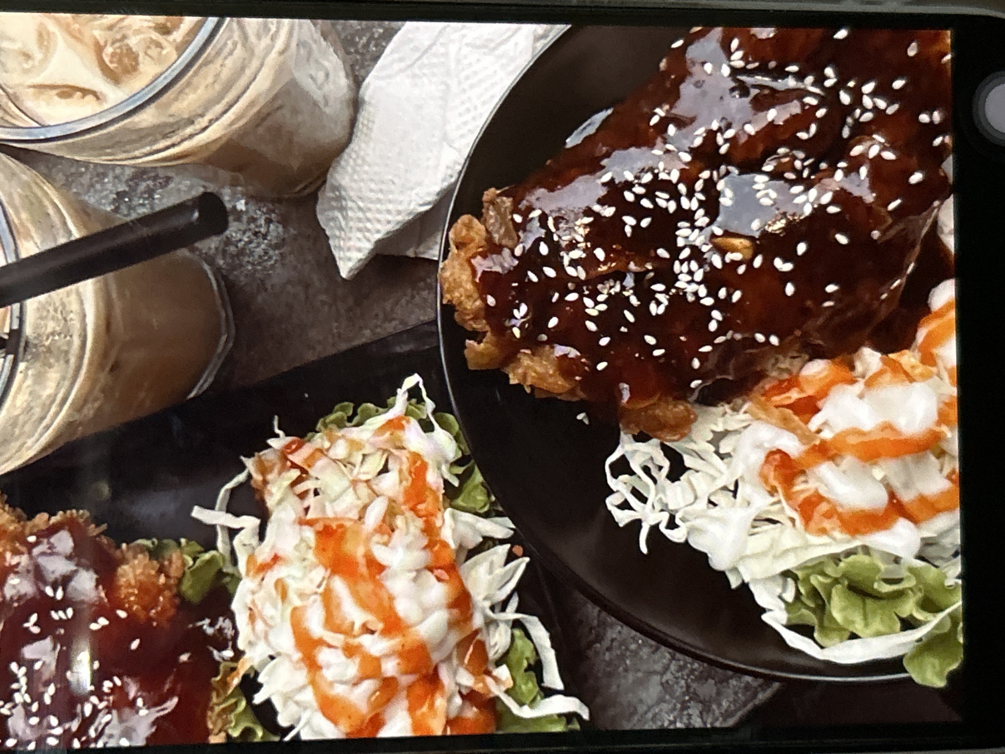 The image shows two plates of food and two glasses of iced coffee on a dark, textured surface. The food on the left plate consists of what appears to be fried chicken or pork, covered in a dark glaze and sprinkled with white sesame seeds. It is served with a side of shredded cabbage and lettuce, drizzled with a white sauce and an orange sauce. The plate on the right also contains a portion of the glazed meat with sesame seeds, served with shredded cabbage and lettuce, also dressed with sauces.
The two glasses contain iced coffee, with visible ice cubes and a creamy, light brown liquid. One glass has a black straw. A white paper napkin is visible between the food and the drinks.
The scene is likely from a cafe or restaurant, given the presentation of the food and drinks. The lighting suggests an indoor setting. The image appears to be a close-up shot, possibly taken with a mobile phone, with a slight blur in some areas. The overall impression is of a meal being served.
While the prompt states the image is from Depok, Indonesia, there are no visual cues within the image itself that confirm this location. This information is external to the visual content.