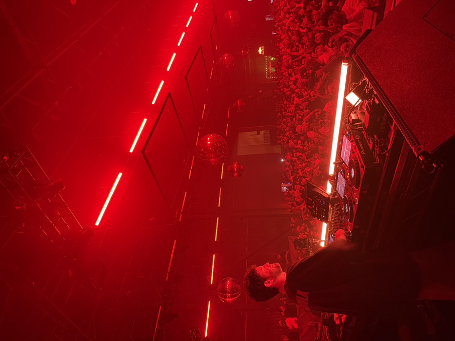 The image depicts an indoor electronic music event taking place in Los Angeles, United States of America. A male disc jockey, seen in profile from the left, is positioned behind a professional DJ setup. He wears a dark short-sleeved garment with light-colored lettering, possibly "SOLA," on the back. His hands are near the controls of Pioneer DJ equipment, which includes a mixer and two CDJ players illuminated by linear red lights integrated into the performance platform.
In the mid-ground and background, a large, densely packed crowd of individuals faces the stage, predominantly obscured by the deep red ambient lighting. Numerous hands are visible raised above the crowd. The venue's ceiling is dark, featuring multiple linear red light fixtures arranged horizontally, interspersed with several large, spherical disco balls that reflect the pervasive red illumination. A blurred red and white logo, possibly for "Red Bull," is faintly discernible in the far background above the audience. The entire scene is immersed in a strong red light emanating from various sources.