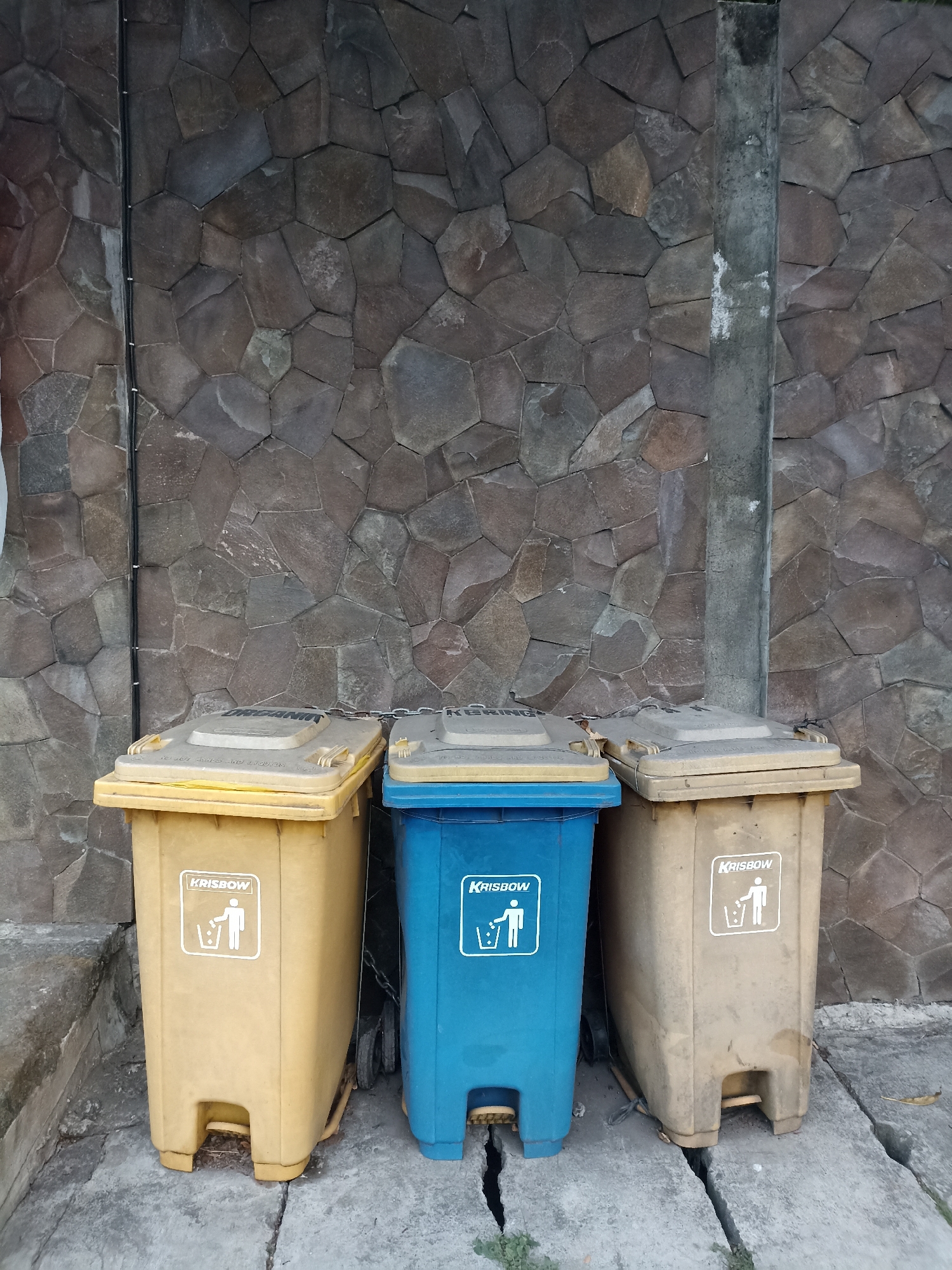 Three trash bins are lined up against a stone wall in Mojopanggung, Indonesia. The bins are yellow, blue, and tan. Each bin has a lid and a symbol of a person throwing trash into a bin. The stone wall is made of irregular, flat stones in shades of brown and gray. A concrete pillar is visible to the right of the bins. The ground in front of the bins is concrete and cracked.