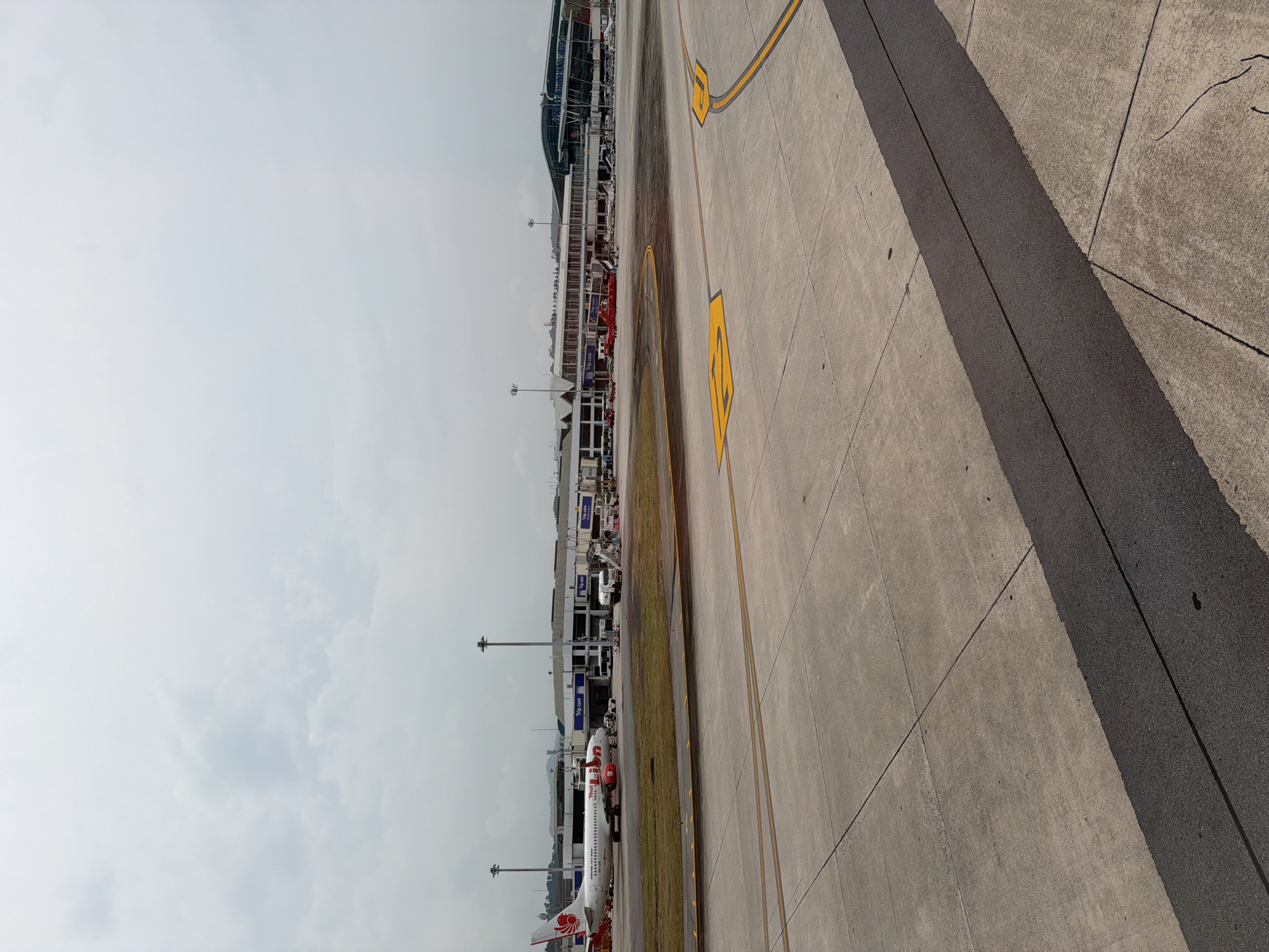 This is an outdoor shot of an airport setting in Tambon Mai Khao, Thailand. An airplane is parked next to a terminal building that features "Trip.com" signage. The airport's concrete tarmac is prominent, displaying yellow markings and expansion joints. The sky is overcast.