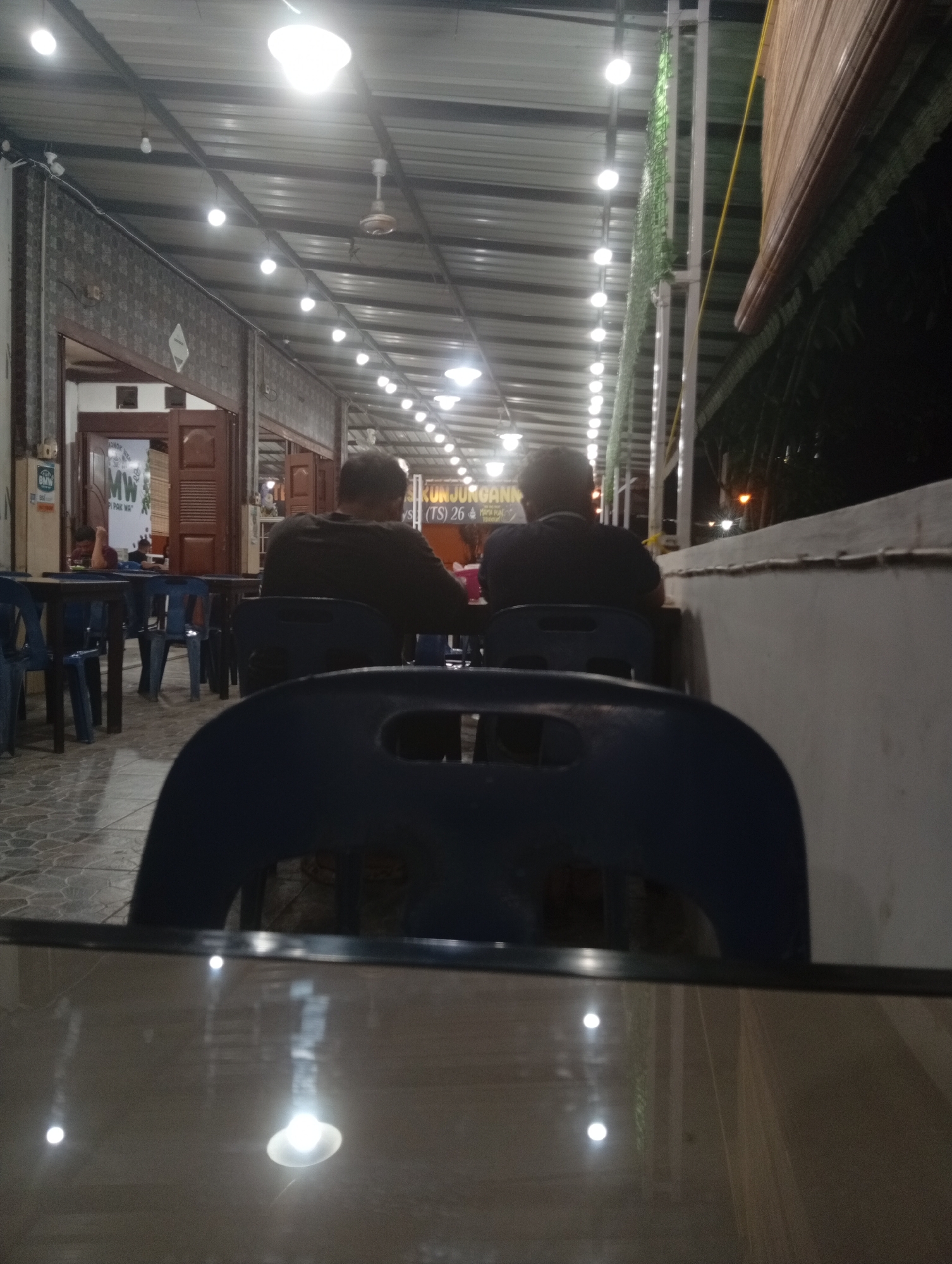 The image depicts an outdoor seating area of a restaurant at night, likely in Lhokseumawe, Indonesia, indicated by the sign that reads "KUNJUNGA... (TS) 26". Two men are seated at a table, seen from behind. The man on the left is wearing a dark t-shirt, while the man on the right is wearing a dark long-sleeved shirt. They are seated at a table in front of them, with blue chairs surrounding it. The restaurant appears to be covered by a corrugated metal roof supported by white pillars, with numerous string lights illuminating the space. In the background, there are doorways leading into the restaurant, with some signage visible, including "MW" and "PAK WAY". To the right of the seating area, there is a wall and some vegetation. The foreground shows the glossy surface of a table, reflecting the overhead lights. The overall impression is of a casual dining establishment at night.