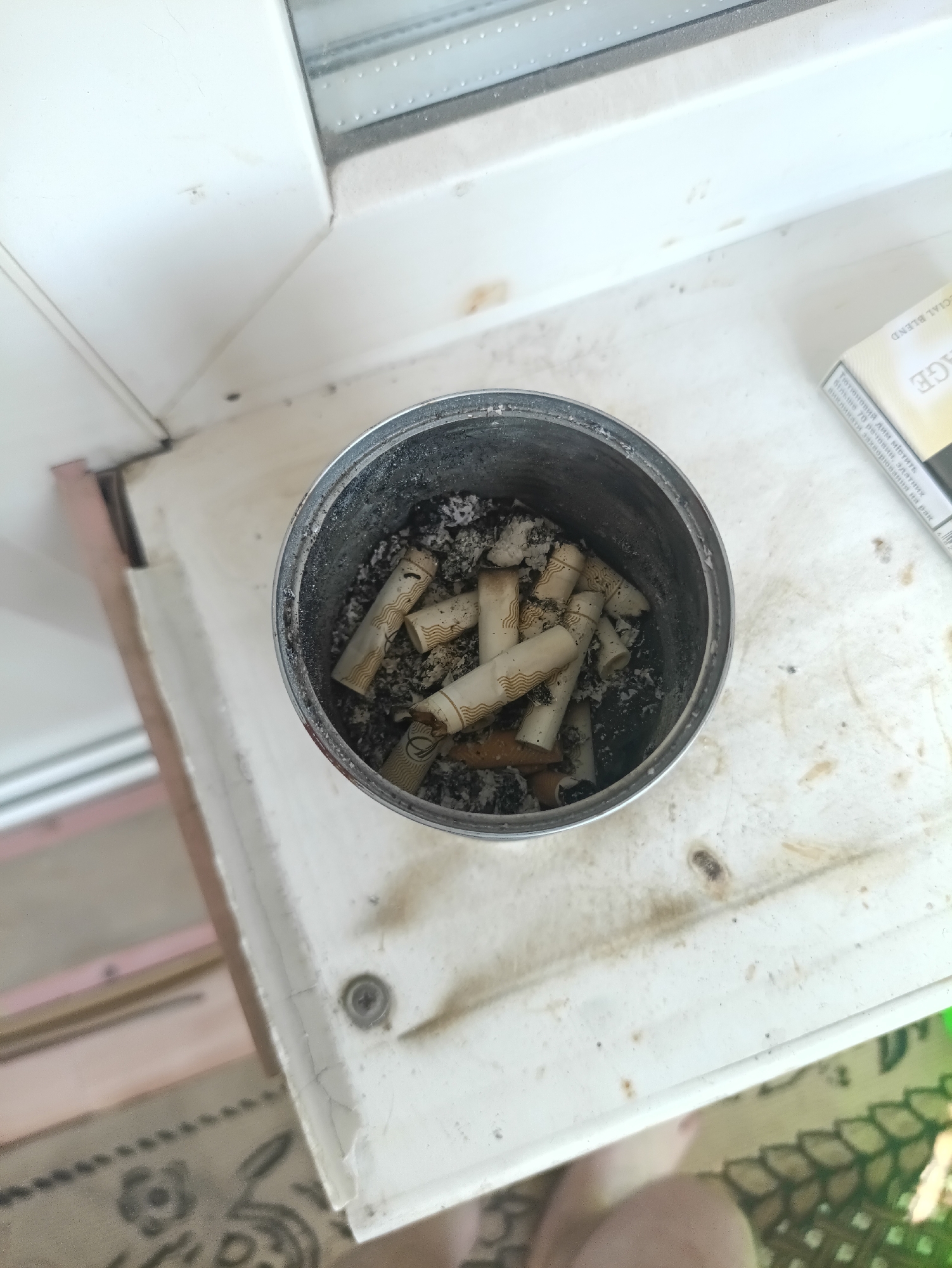 The image shows an ashtray filled with cigarette butts on a windowsill. The ashtray is a metal can. The windowsill is white and appears to be made of plastic or painted wood. There is a cigarette pack with the text "CIAL BLEND" and some text in Ukrainian visible on the right side of the image, indicating the location context is Kamianets-Podilskyi, Ukraine. The overall scene suggests a moment of smoking, likely indoors. The lighting suggests it is daytime. There are no people fully visible in the image, but a portion of a leg and foot are present at the bottom. The scene appears somewhat neglected, with dust and grime visible on the windowsill.