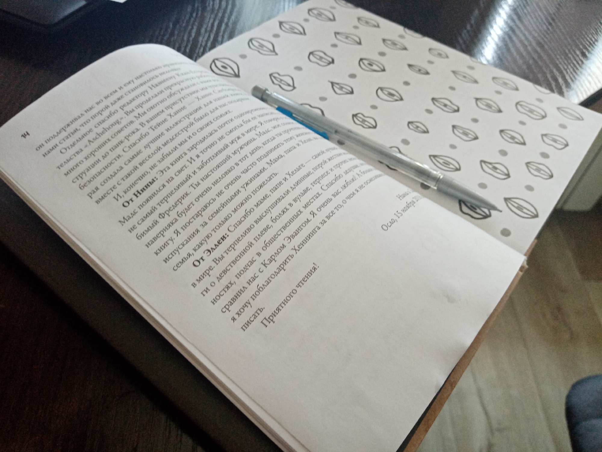 The image shows an open book lying on a dark surface, possibly a desk or table. A silver pen rests on the pages of the book. The book has black and white patterned end papers consisting of what appear to be abstract eye shapes and dots. The book appears to be a dedication of sorts, in Russian, featuring acknowledgements to various people, including editors and family members. The location in the city of Elektrostal, Russia is not possible to ascertain from the image.