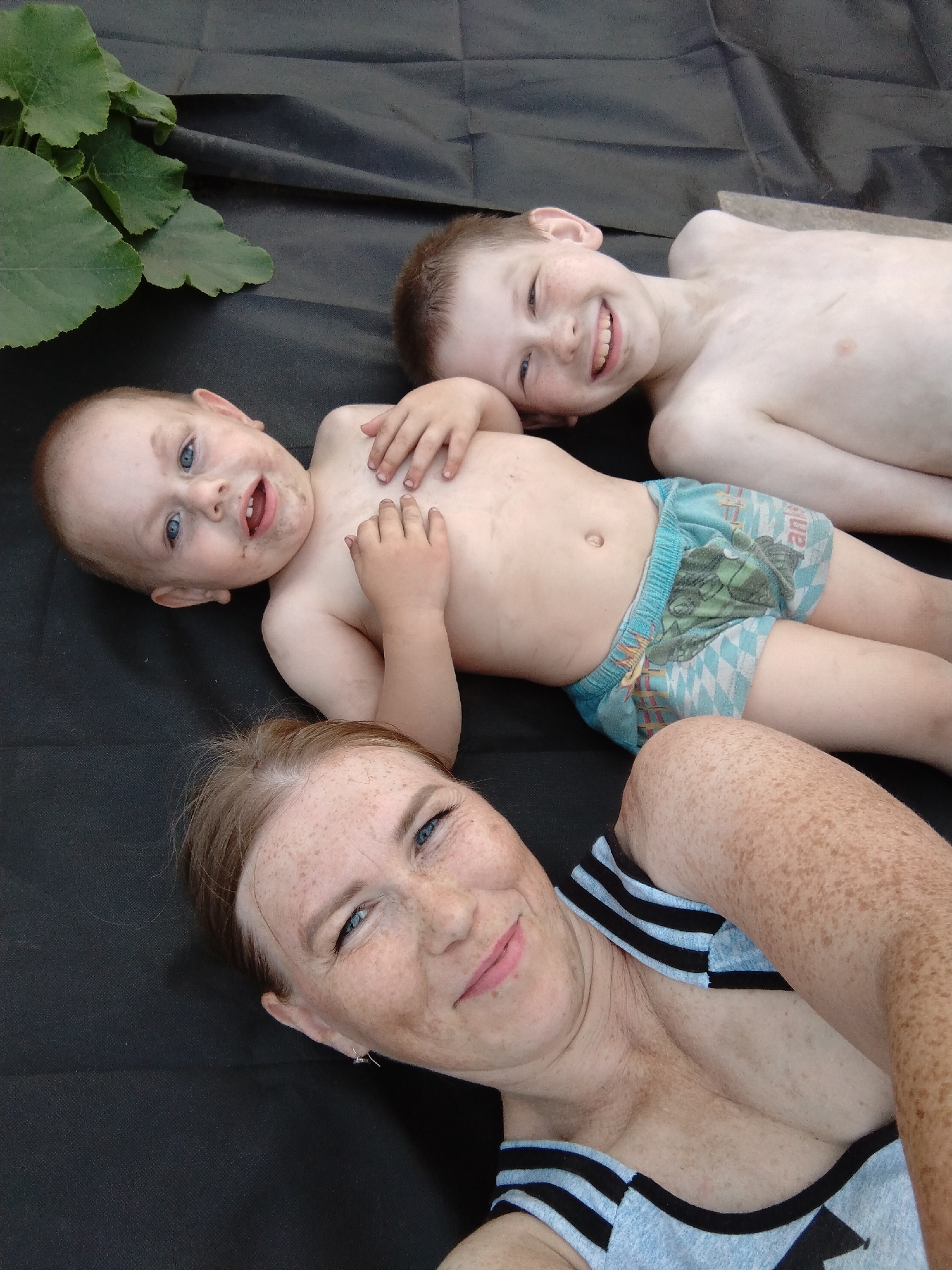 In this image, three people are lying on a dark, textured surface, possibly a blanket or ground cover. The subjects are two young boys and an adult woman. The woman is in the foreground, on the right side of the frame, with her head tilted towards the camera. She has light brown hair with a reddish tint, freckles across her face and arms, and is smiling. She is wearing a light blue top with black stripes along the neckline and straps.
Behind the woman, a younger boy with light hair and freckles is lying on his back, looking up and smiling broadly, with his mouth open in a joyful expression. He is wearing light blue patterned shorts with cartoon characters on them. To his left and slightly behind him, another younger boy, possibly a toddler, is also lying on his back. He has a bald head, blue eyes, and is looking towards the camera with his mouth open, appearing to be speaking or making a sound. His chest and arms are visible, and he has some dirt smudges on his skin.
In the upper left corner of the image, large green leaves from a plant, possibly a pumpkin plant given the context of Komsomolsk-on-Amur, Russia, are visible. The overall scene appears to be outdoors, perhaps in a garden or backyard, on a day with natural lighting. The expressions of the subjects suggest a happy and relaxed moment. There is no visible text in the image.