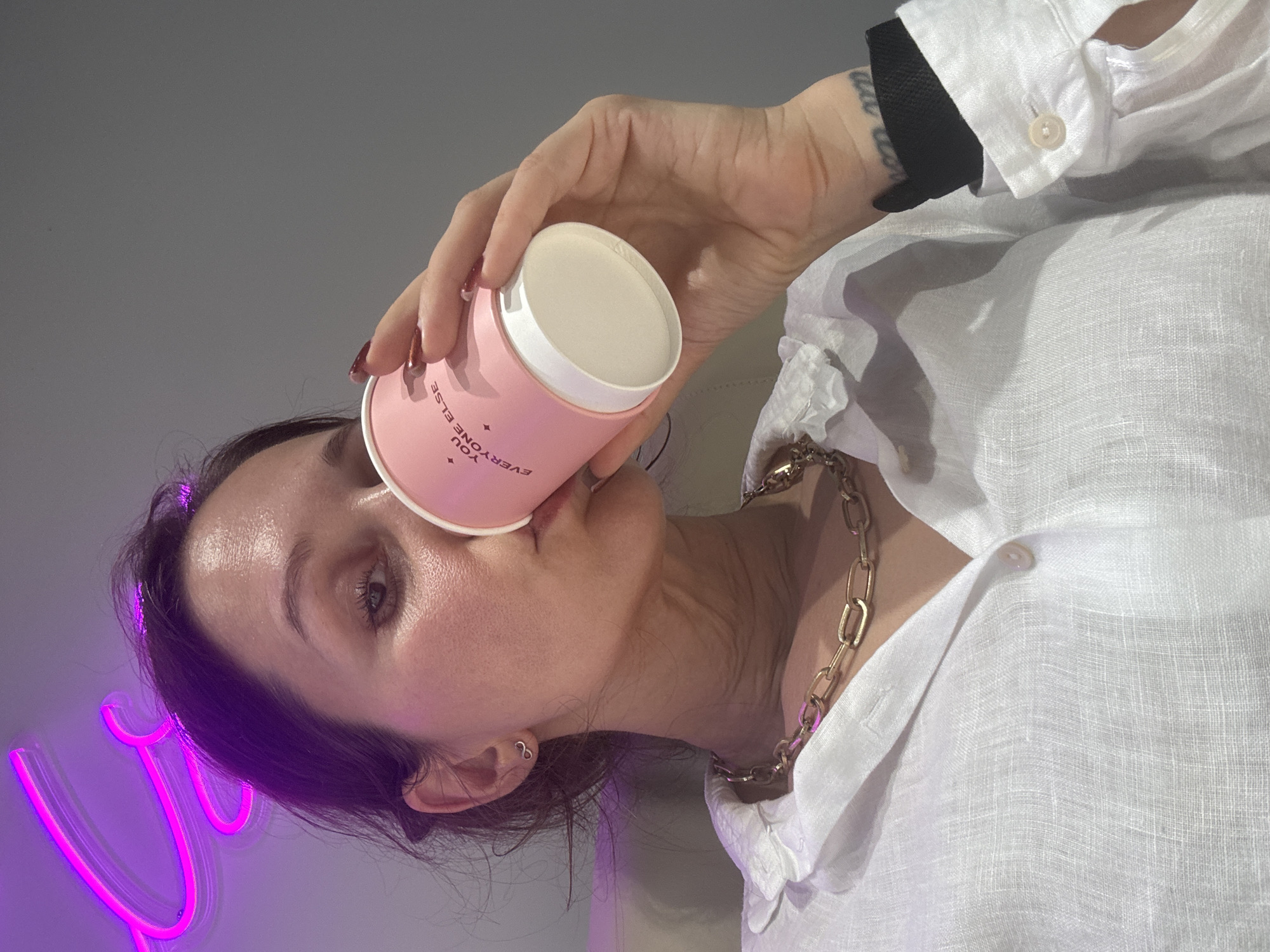 A woman is angled to the right, holding a pink and white cylindrical container labeled "YOU EVERYONE ELSE." She is wearing a white linen shirt with a visible gold chain necklace. Her left wrist has a black wristband and a tattoo. Her makeup is minimal, and her hair is dark. A neon pink light fixture is to her lower left. The background is a plain grey wall. There is no indication that the image was taken in Dubai, United Arab Emirates.