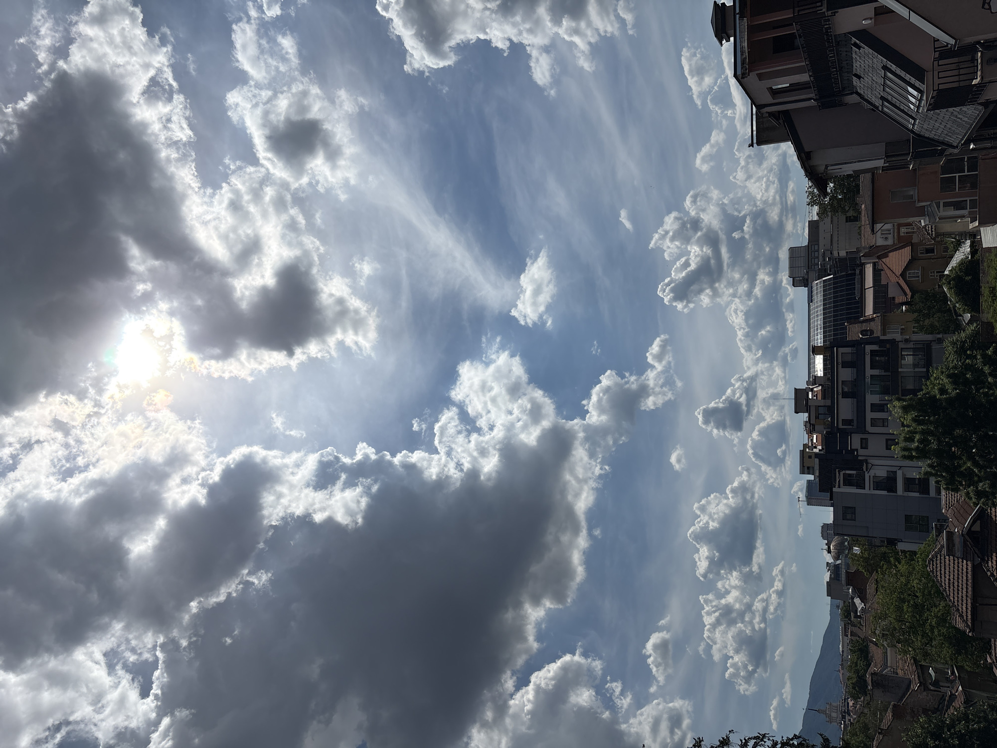 The image shows a city skyline with a dramatic sky filled with clouds. The sun is partially visible through the clouds, casting a bright glow. The buildings in the foreground are a mix of old and new architecture, with tiled roofs and balconies. In the background, a mountain range is visible. The scene appears to be in Sofia, Bulgaria, possibly looking towards Vitosha Mountain. There are no people or actions depicted in the image, and the focus is on the architecture and the sky.