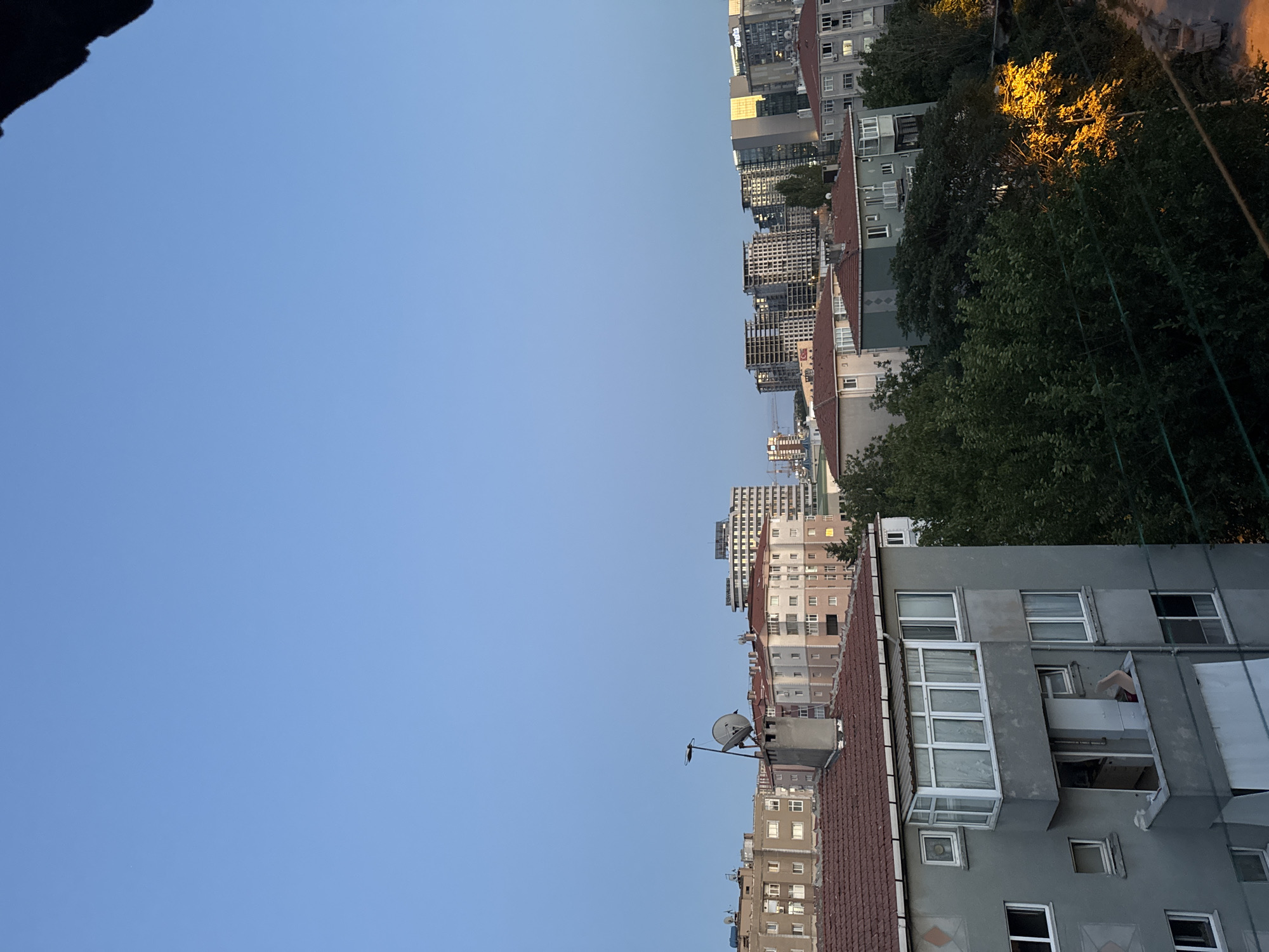 The image shows an outdoor urban scene at dusk in Gungoren, Turkey. The sky is a clear, soft blue, indicating the transition from day to night. In the foreground, a gray, multi-story residential building with a tiled roof is visible. It has several windows and a enclosed balcony. Behind this building, a row of older apartment buildings with reddish-brown tiled roofs stretches across the mid-ground. Some of these buildings have satellite dishes mounted on their roofs. Further in the background, modern, taller buildings, some under construction, dominate the skyline, reflecting the city's ongoing development. Lush green trees are interspersed between the buildings, with some illuminated by a warm, yellow light, possibly from streetlights. A dirt path or road with construction materials is visible on the right side of the image, suggesting ongoing construction or maintenance in the area. The overall impression is of a residential district experiencing a blend of older architecture and new construction, set against a tranquil twilight sky. There are no people or animals visible in the image.