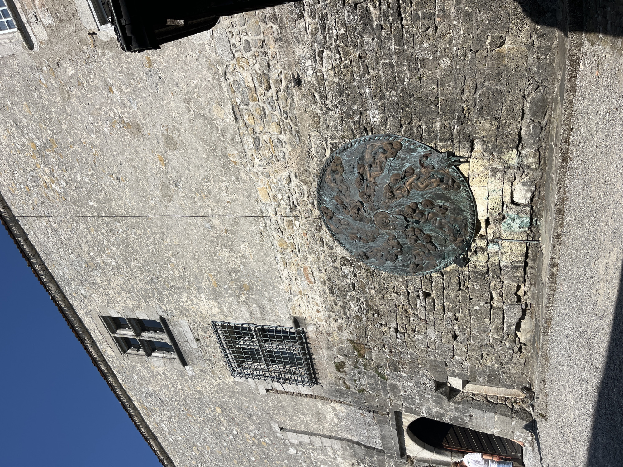 The image shows a section of a weathered stone building, likely a historic structure, in Epagny, Switzerland. The building's facade is constructed of rough-hewn stones of varying sizes and colors, interspersed with patches of stucco or plaster. A large, circular bronze relief sculpture is mounted on the wall, depicting numerous intertwined figures. To the left of the sculpture, an arched doorway is visible, with a wooden door ajar. A person, dressed in a white shirt and blue shorts, is standing partially in the doorway, facing away from the camera. Above the doorway, a rectangular window with a wrought-iron grate is set into the wall. Further up the wall, another, more elaborate window with stone mullions is visible. The ground in front of the building is covered in gravel. The sky above is a clear, bright blue, indicating a sunny day. A dark shadow falls from the right side of the image, suggesting the presence of another structure or overhang.