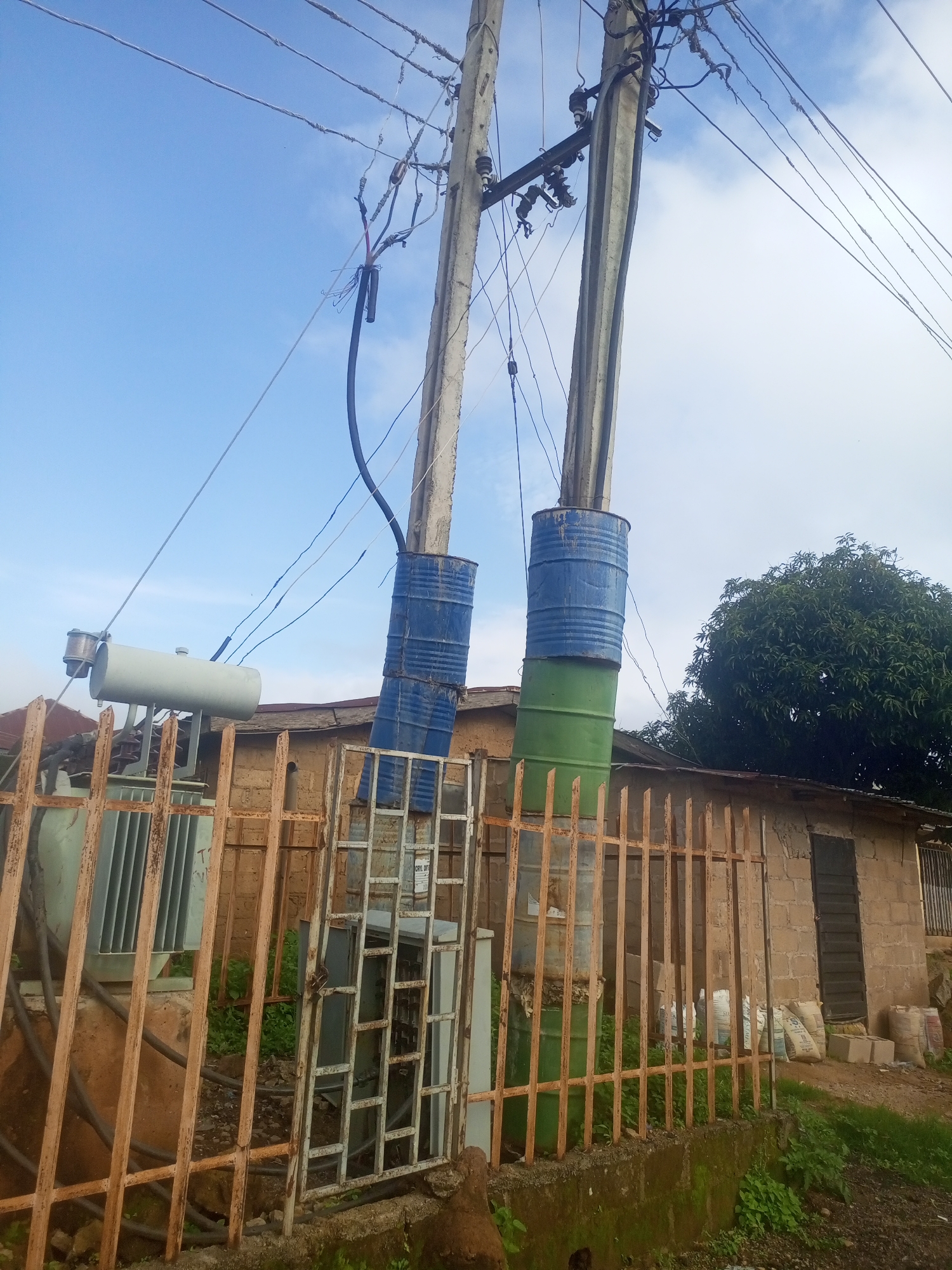 In the city of Jos, Nigeria, a scene of electrical infrastructure is depicted. Two concrete utility poles stand, one with a blue drum fitted around its lower section and another with two stacked drums, blue on top and green on the bottom. These drums appear to be repurposed to extend the height of the poles. Overhead power lines crisscross the sky, connecting to insulators on the poles.
At the base of the poles and adjacent to a building, an electrical transformer is visible. It is a light green, rectangular unit with ribbed sides and appears to be connected to the overhead lines by thick black cables. A rusty, orange metal fence runs horizontally across the foreground, partially obscuring the transformer and the building.
The building itself is constructed from rough-hewn, light-colored blocks. It has a corrugated metal roof and a dark, panelled door. Bags of what appear to be cement or building materials are stacked near the entrance of the building. A lush green tree with a wide canopy is situated behind the building. The ground is covered with a mix of dirt, gravel, and patches of green vegetation. The sky above is a bright blue with scattered white clouds, suggesting daytime.