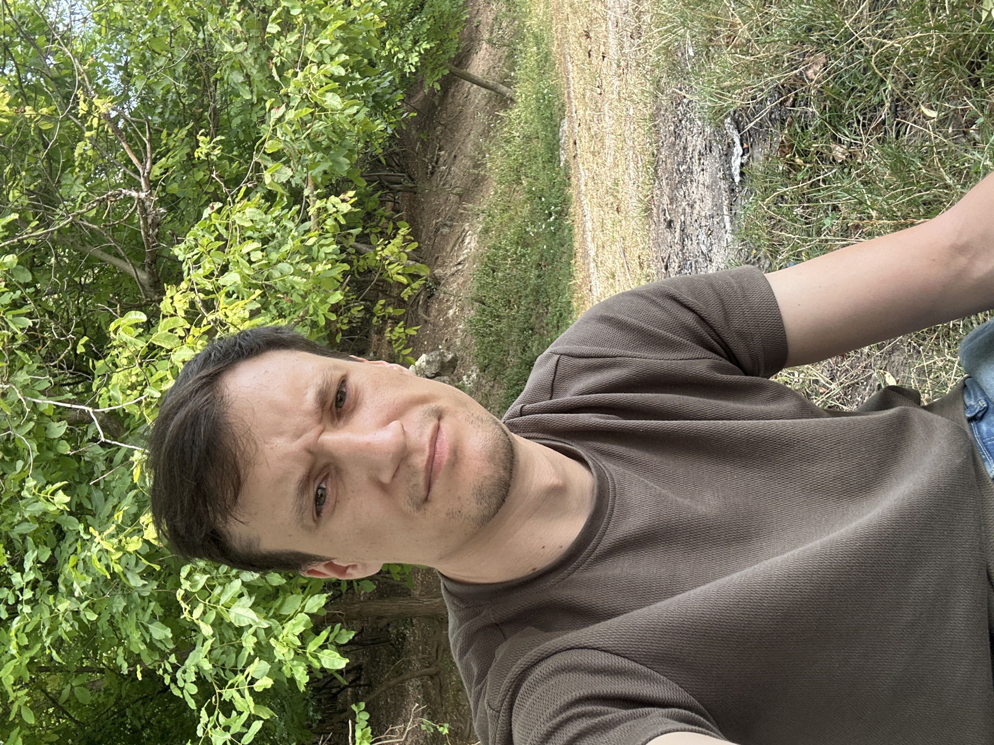 A man with dark hair and stubble is captured in a selfie outdoors, possibly in Makiv, Ukraine, with a backdrop of lush green trees and dry grass. He is wearing a brown t-shirt and blue jeans. His expression is neutral, and he is looking directly at the camera. The lighting suggests it is daytime, with the sun casting soft shadows. The environment appears natural and somewhat unkempt, with visible dirt patches and scattered vegetation.