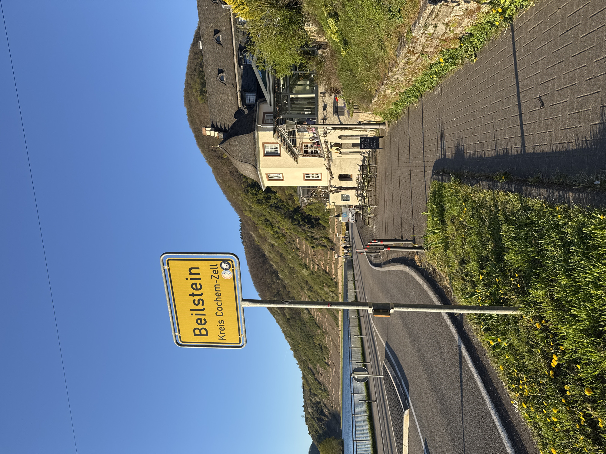 The image captures a roadside scene in Beilstein, Germany. A yellow signpost with "Beilstein Kreis Cochem-Zell" is prominently displayed against a clear blue sky. A road curves along the riverbank with a safety barrier. On the other side of the road, a building sits nestled against a hillside, with visible architectural details like windows, a roof with dormers, and a partially visible exterior staircase. A paved path runs alongside the building. Vegetation, including grass and dandelions, grows on either side of the path. The water of the river is partially visible.