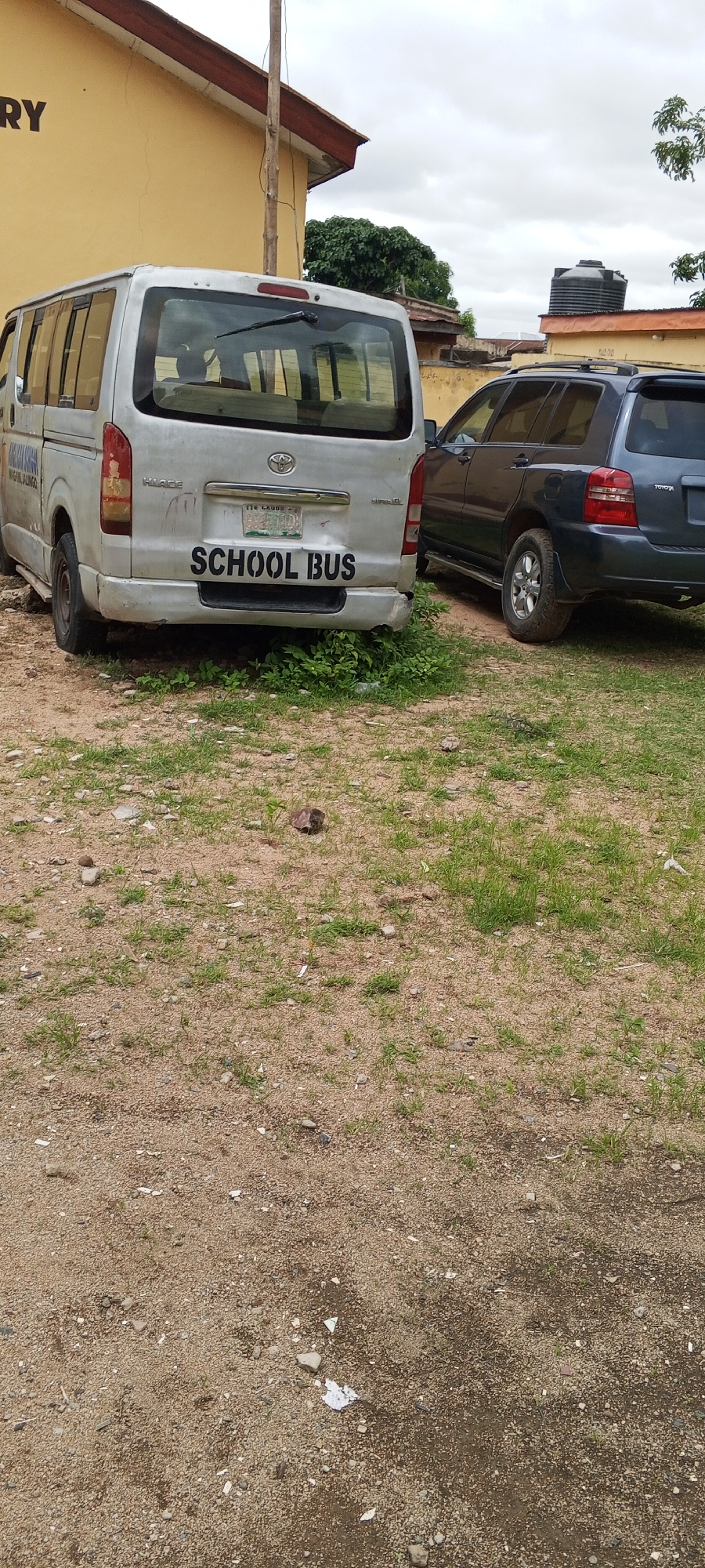 A silver Toyota Hiace van, identified by the text "SCHOOL BUS" stenciled in black on its rear bumper, is parked on a gravel and grass lot. The van's rear window is visible, as is a portion of a dark blue Toyota Highlander SUV parked to its right. The building behind the vehicles is painted yellow and has a brown roof. In the background, a black water tank sits atop a low building. The sky is overcast with clouds. The scene appears to be in Jalingo, Nigeria, suggested by the license plate on the van, which reads "LAGOS 967 1AO1".