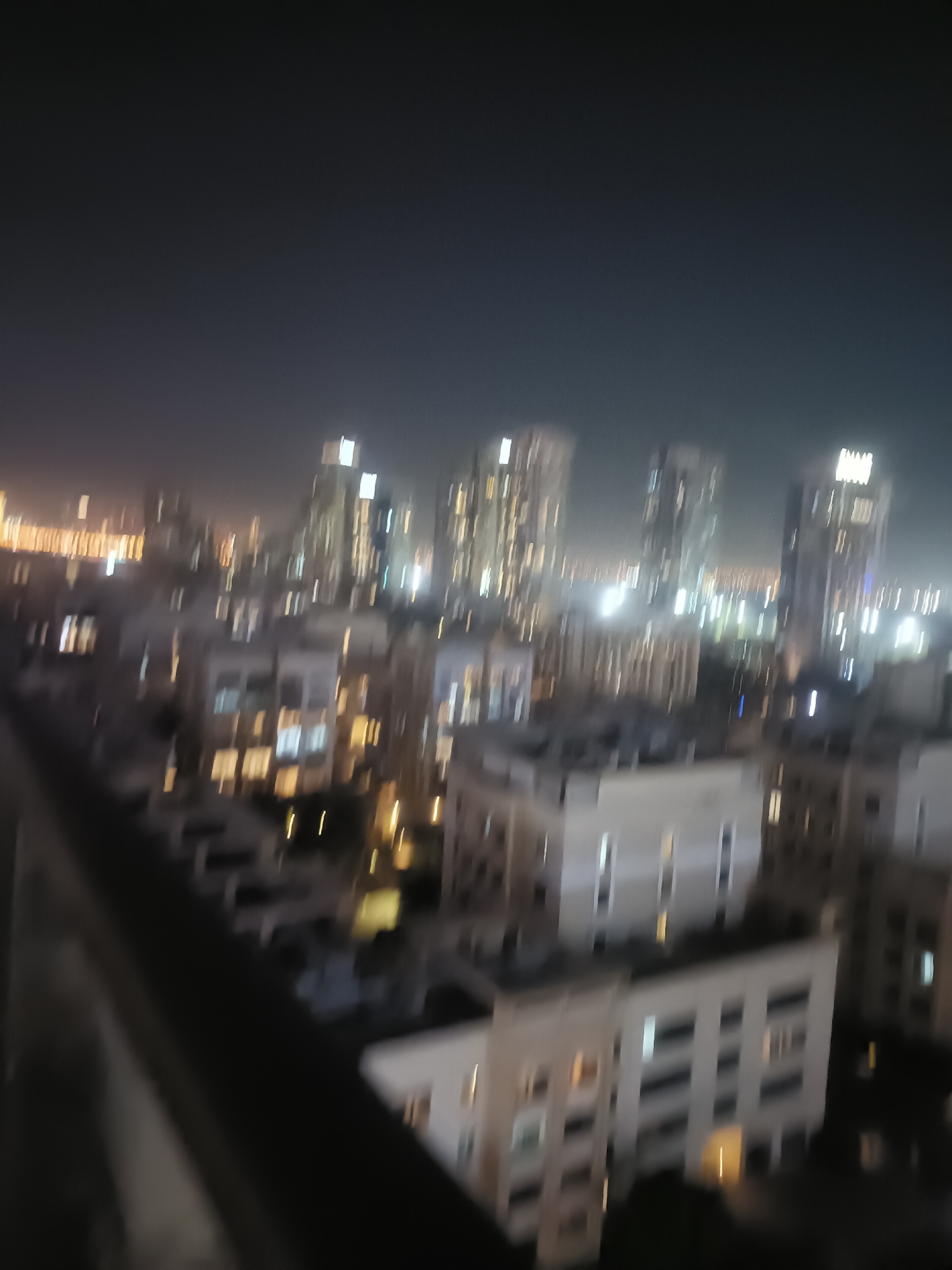 This is a blurry, night-time, aerial view of a cityscape in Dubai, United Arab Emirates. The image shows a cluster of tall buildings, illuminated by numerous lights. The lights suggest a dense urban environment, likely a business or residential district. In the foreground, a dark railing or balcony suggests the viewpoint is from a high-rise building. The overall impression is of a vibrant, well-lit city at night.