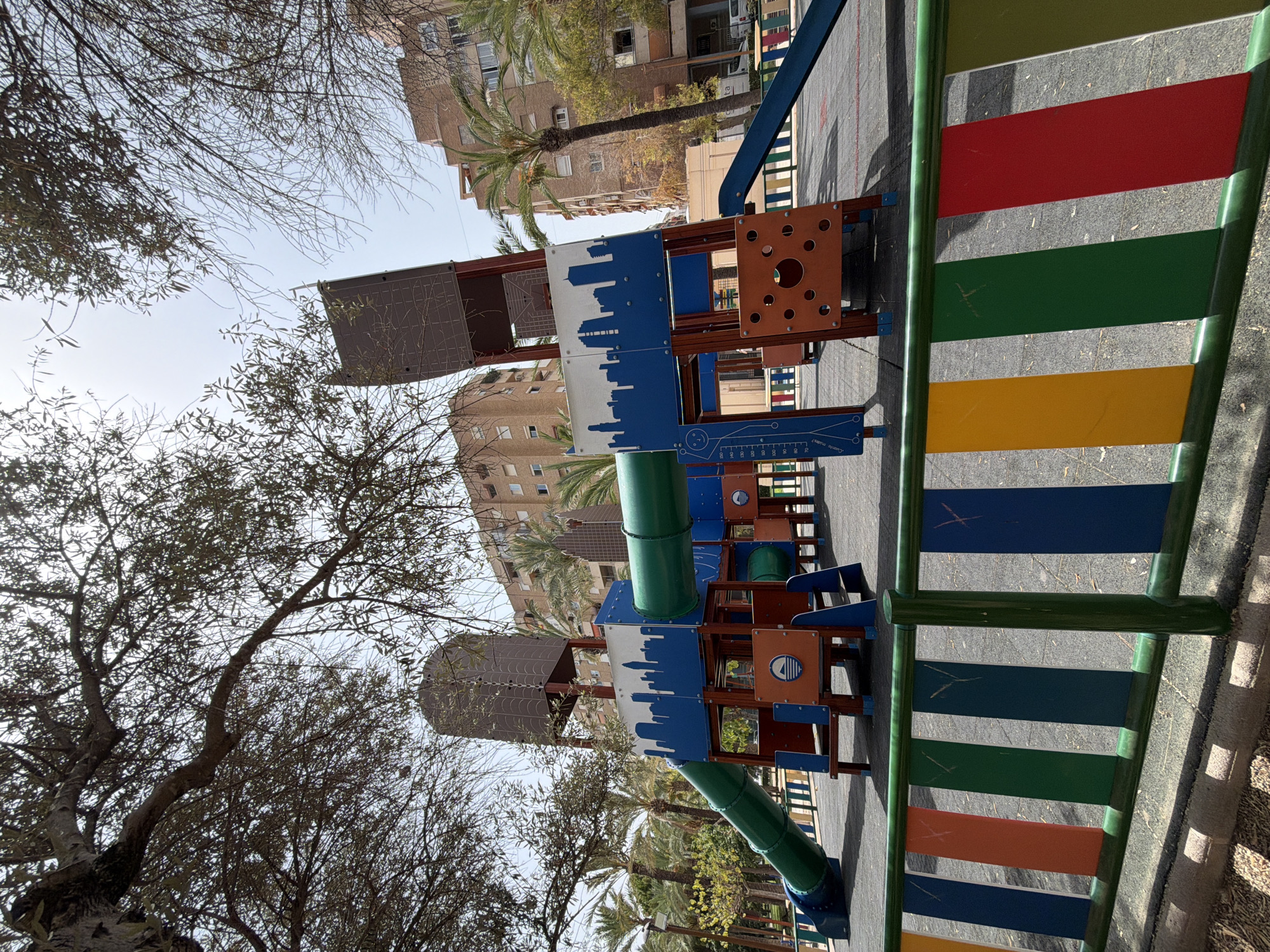 The image shows a brightly colored children's playground in Benidorm, Spain. The playground features a large, multi-level structure with slides, tubes, and platforms. A green tube slide descends from the left side of the structure, and a blue slide is visible on the right. The structure is adorned with blue panels depicting a white cityscape. Orange panels with circular cutouts are also present. The ground of the playground is a dark, textured surface. A fence with vertical slats in various colors, including green, blue, orange, and red, separates the playground area from the surrounding environment. In the background, there are tall apartment buildings and palm trees, characteristic of the urban landscape of Benidorm. The sky is visible above, with branches of trees framing the top of the image. No people are present in the image.