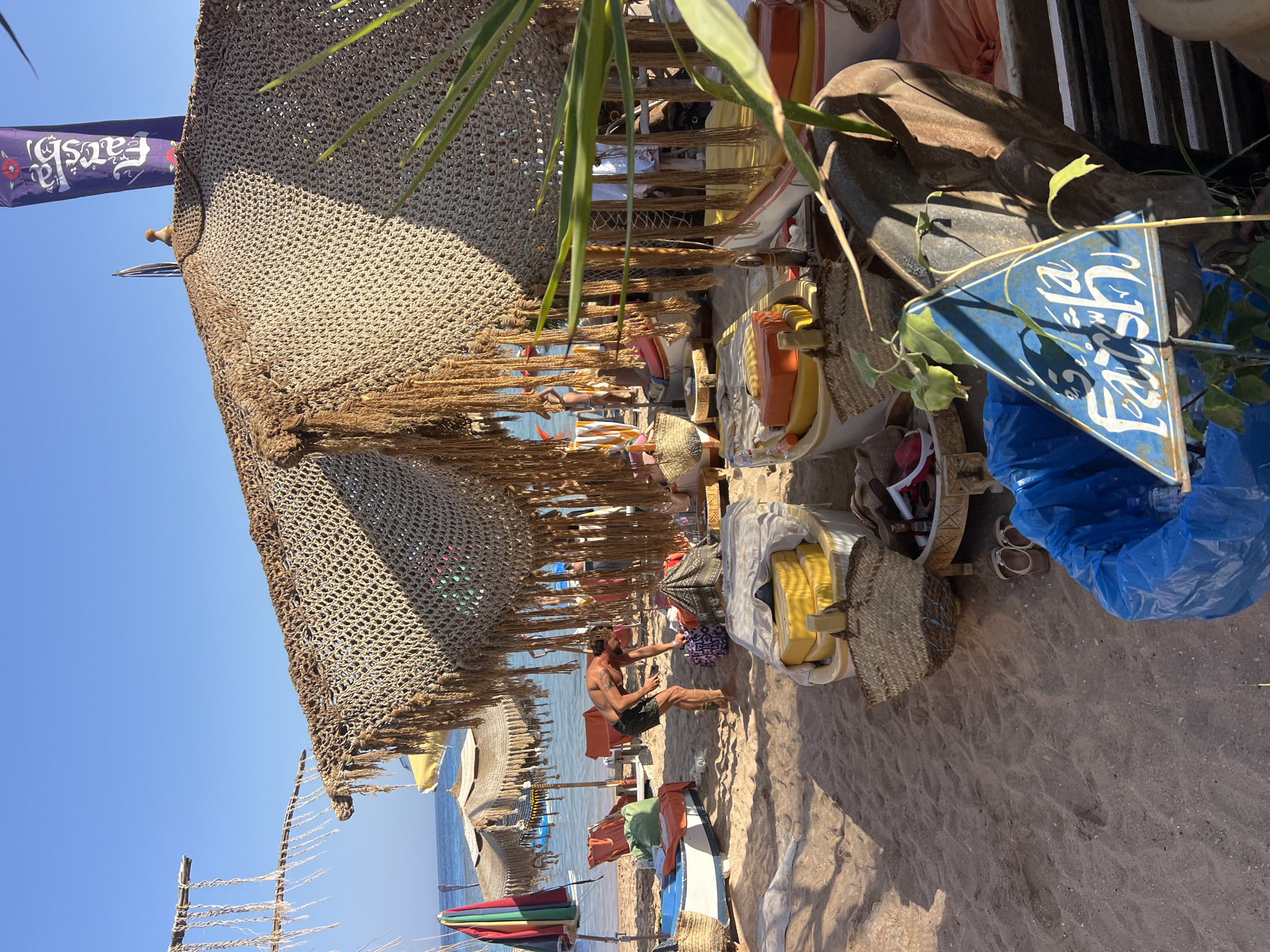 The image depicts a beach scene in Sharm El Sheikh, Egypt, on a sunny day. In the foreground, a blue trash bag is visible, partially filled with items, with a blue triangular sign with Arabic script and the word "Faith" in English attached to a plant. Behind this, a rustic thatched umbrella provides shade over several boats that have been converted into seating areas. These boats are adorned with yellow and orange cushions. A man in black shorts is standing in the sand, leaning over and interacting with a patterned bag. Several beach chairs and other umbrellas are scattered across the sandy beach, leading to the clear blue water in the background. The overall atmosphere suggests a relaxed beachside establishment.