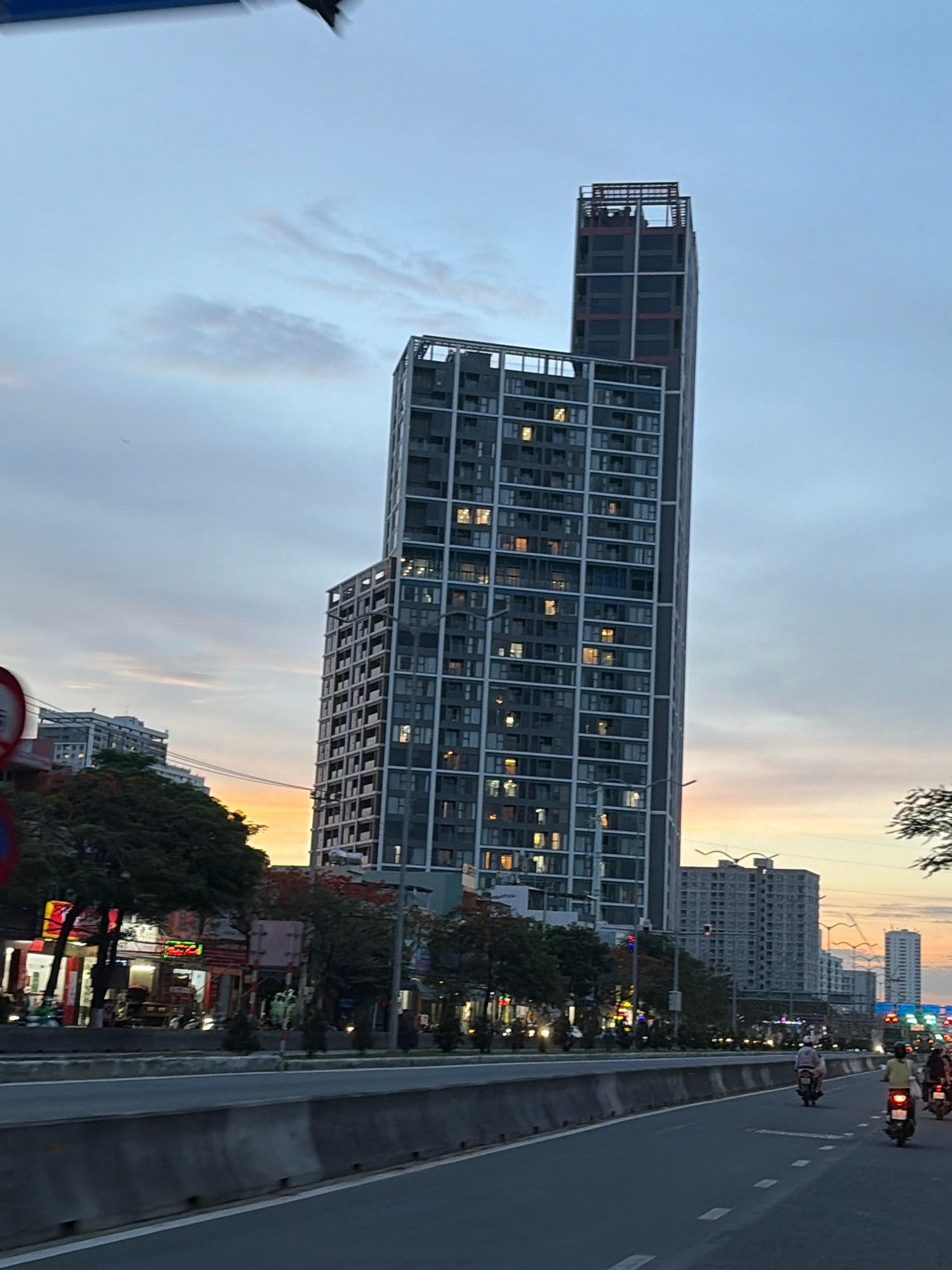 This image depicts a street scene in Da Nang, Vietnam, during what appears to be dusk or early evening. The central focus is a very tall, modern apartment building with a dark facade and numerous windows, many of which are lit. To the left of this main building, a slightly shorter, similarly styled building is visible. Further in the background, other buildings of varying heights are present, some appearing to be under construction. The sky is a gradient of pale blue, with hints of orange and pink near the horizon, suggesting a sunset.
In the foreground, a road runs from left to right, divided by concrete barriers. Several motorbikes are traveling along the road, with riders visible on some. On the left side of the road, there are storefronts with illuminated signs, and trees with lush foliage. Traffic lights are visible in the distance. The overall impression is of a busy urban environment transitioning into the evening.