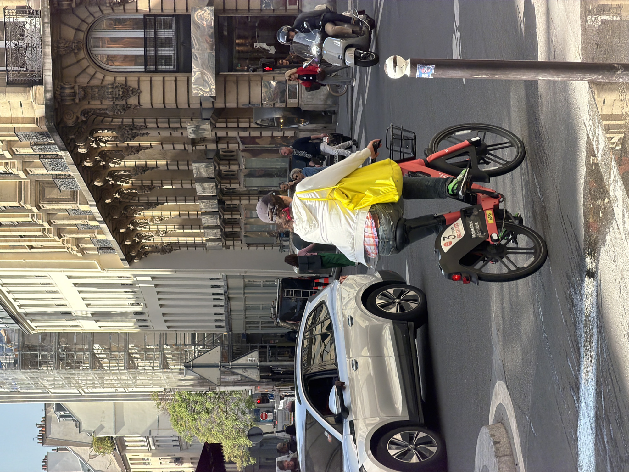 The image depicts a street scene in Paris, France. A person is riding a red and black bike. A silver car is driving down the street. Other people are walking on the sidewalk. Buildings with architectural details line the street. A scooter and bicycle are parked near a lamp post. The street surface appears wet.