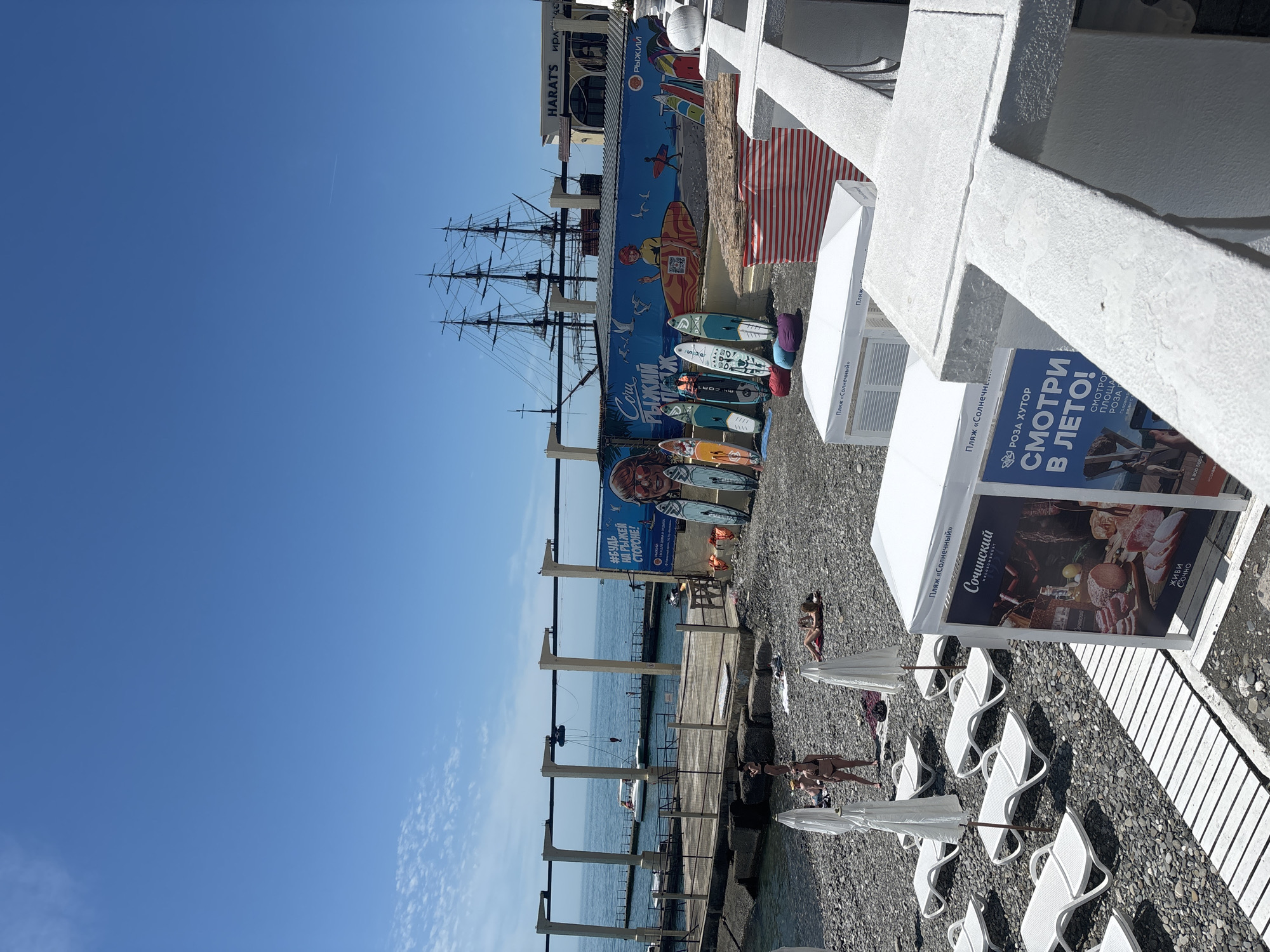 The image depicts a lively beach scene in Sochi, Russia, on a bright, sunny day with a clear blue sky. In the foreground, a pebbly beach is furnished with numerous white lounge chairs and umbrellas, where several people, some in swimwear, are seen relaxing or walking.
Towards the midground, the calm, blue sea stretches out, with a wooden pier extending into the water. Along the shoreline, a long retaining wall is painted with colorful murals featuring a large sailing ship, surfers, and beach-related graphics. Various paddleboards are propped against this wall. Behind the mural, architectural details of buildings are visible, including one with the text "HARAT'S".
Notable details include the brilliant sunlight, the absence of clouds, and the clear distinction between the sea and sky. Visible text on signs and banners provides location cues and advertisements. `Пляж «Солнечный»` (Sunny Beach) is repeatedly seen on white tents, identifying the beach area. A large blue banner prominently advertises `РОЗА ХУТОР СМОТРИ В ЛЕТО!` (Roza Khutor Look in Summer!), referencing a well-known resort. Other texts include `#БУДЬ НА РЫЖЕЙ СТОРОНЕ!` (Be on the Red/Ginger Side!), `Coru РЫЖИЙ Ж` (Sochi Red), and `Сочинский` (Sochi) with the slogan `живи Сочно` (Live Juicily/Vibrantly). The scene captures a typical summer resort ambiance with leisure and outdoor activities.