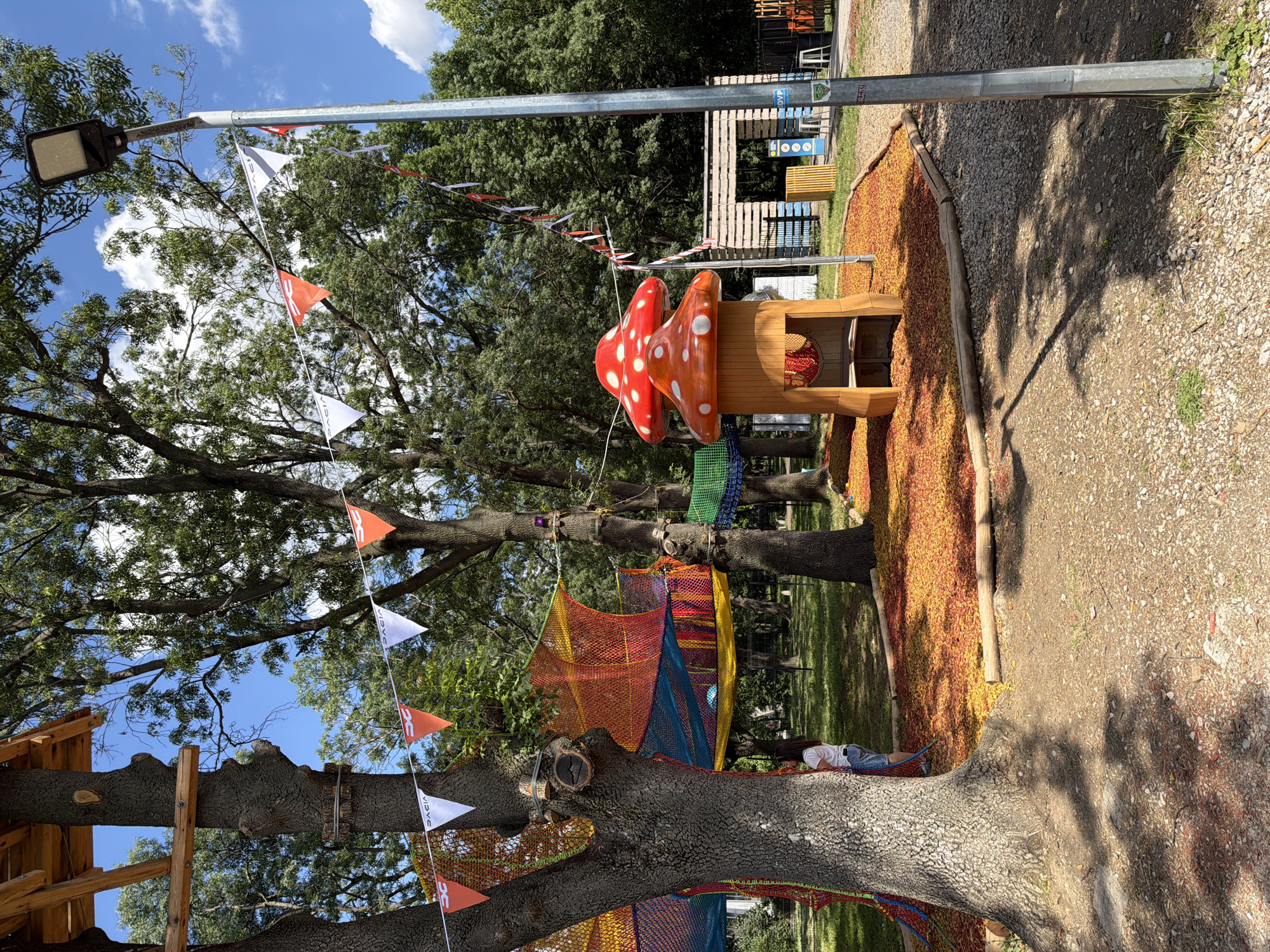 The image captures a children's adventure park in Sofia, Bulgaria, on a bright, sunny day. A large, textured tree trunk dominates the left side of the frame, partially obscuring a colorful, netted climbing structure suspended between trees. This structure features bright red, orange, yellow, and blue netting. A young girl, wearing a white t-shirt and blue denim shorts, stands partially hidden behind the tree, looking towards the climbing area.
In the foreground, a whimsical mushroom-shaped playhouse with red and white spots stands on a bed of red and yellow wood chips. A string of triangular pennants, some orange and white with a "DACIA" logo, and others red, white, and black, stretches across the scene, attached to trees and a lamppost. The lamppost, a tall, metallic pole with a modern light fixture, is positioned on the right side of the image.
Behind the playhouse, a rustic wooden structure made of pallets serves as an entrance or shelter, with signage and seating visible nearby. The ground is a mix of gravel and dirt, with scattered wood chips and a few small plants. The background is filled with the lush green foliage of mature trees, and the sky above is a clear blue with scattered white clouds. The overall atmosphere is playful and inviting, designed for outdoor recreation.