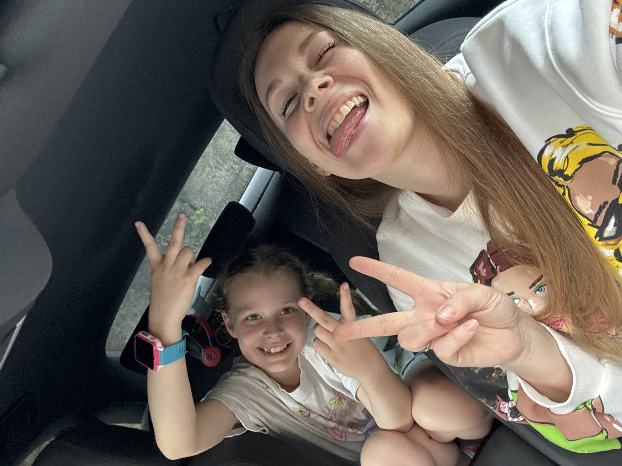 A selfie taken inside a car shows two smiling females making the peace sign gesture. The older female is sticking out her tongue. The younger female is wearing a blue smartwatch and headphones. The location is likely in the city of Budva, Montenegro.