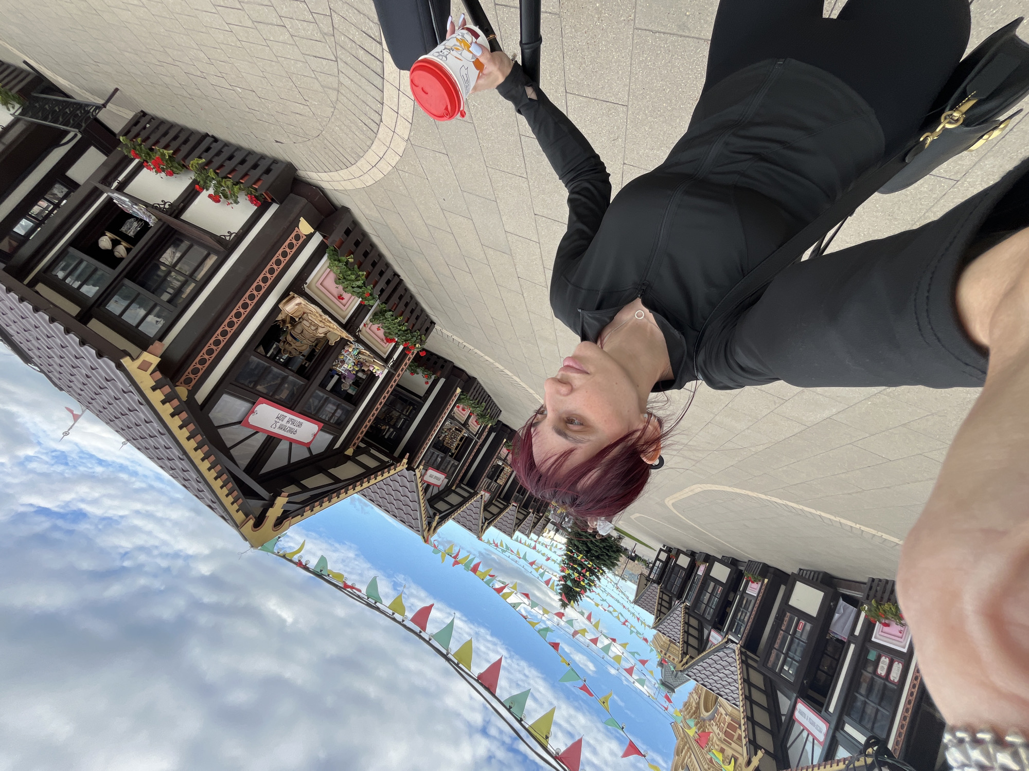 A woman with short, dark red hair is taking a selfie while holding a red and white paper cup and pushing a stroller. She is wearing a black zip-up jacket over a black shirt and black leggings. The background shows a row of small, Bavarian-style market stalls with brown roofs and white trim, decorated with red flowers. Colorful triangular flags are strung between the stalls, and a decorated Christmas tree is visible in the distance. The ground is paved with light-colored tiles. The scene is located at the Nizhny Novgorod Christmas Market.