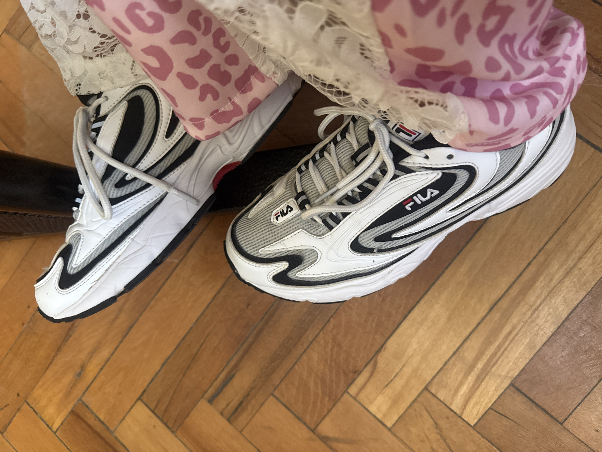 The image shows a close-up of two FILA brand athletic shoes on a wooden parquet floor. The shoe on the left is predominantly white with black stripes and a black outsole. The shoe on the right is predominantly white and gray with black accents and a white outsole. Both shoes have white laces and the FILA logo is visible on the side of each shoe. The shoes are partially obscured by clothing; the left shoe is next to pink fabric with animal print, while the right shoe is next to white lace fabric and pink fabric. No people are visible. The location in the city of Elhovo, Bulgaria is not evident from the image.