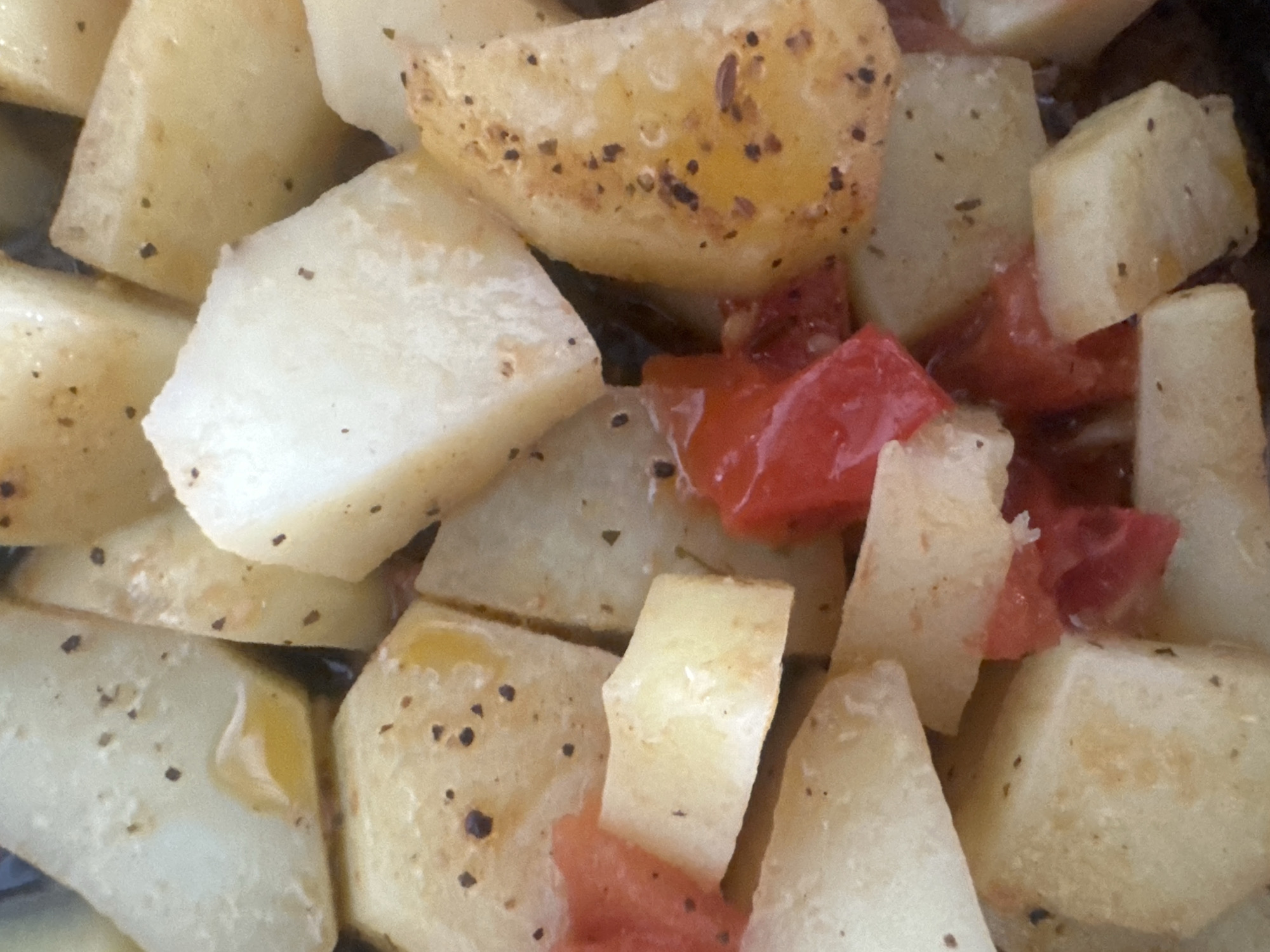 This is a close-up, overhead shot of a dish of roasted potatoes and tomatoes. The potatoes are cut into bite-sized cubes and are lightly seasoned with black pepper and herbs. Some of the potatoes show a golden-brown sear, indicating they have been roasted to a tender, slightly crispy texture. Interspersed with the potatoes are chunks of bright red tomato, which appear to have softened and released some of their juices during the cooking process. A few drops of oil are visible, adding a sheen to the dish. The overall impression is of a hearty, home-cooked meal. The context of Zvezdara Urban Municipality in Serbia suggests this dish might be part of a traditional Serbian meal, perhaps served as a side dish or a light main course. There is no visible text in the image, nor are there any indications of time of day, weather, or specific emotions.