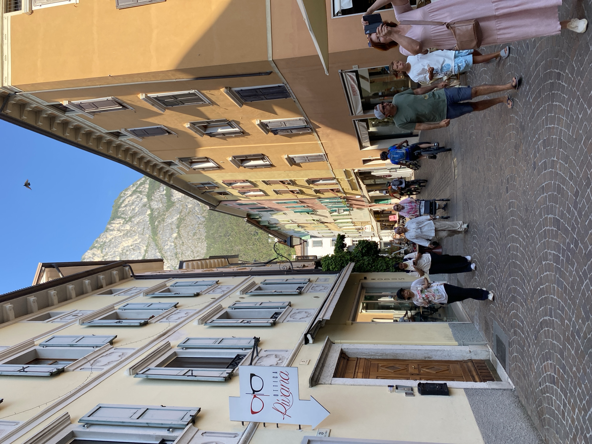 The image depicts a street scene in Riva del Garda, Italy. The street is paved with cobblestones and lined with multi-story buildings with colorful facades, primarily yellow and orange. A prominent sign for "OTTICA Rivana" with eyeglasses imagery is visible on the left. Several people are walking and cycling along the street. On the left, a woman in a pink patterned dress walks with a walker. Further down the street, two young women, one wearing a white t-shirt and the other a white t-shirt and black pants, are walking. In the center, a man in a green t-shirt and blue shorts is walking while a young boy in a white t-shirt and blue shorts walks beside him. A woman in a pink dress is taking a photo with her phone on the right side of the image. Several cyclists are also present in the background. The street opens up to a view of a steep, rocky mountain in the distance under a clear blue sky. The buildings have traditional shutters on the windows, and some have awnings over shopfronts.