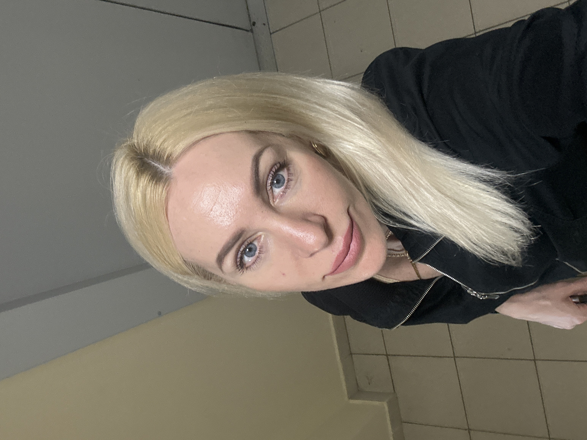 A blonde woman with blue eyes is facing the camera. She is wearing a black jacket. The background consists of beige tiled walls and floor. The location is unknown.