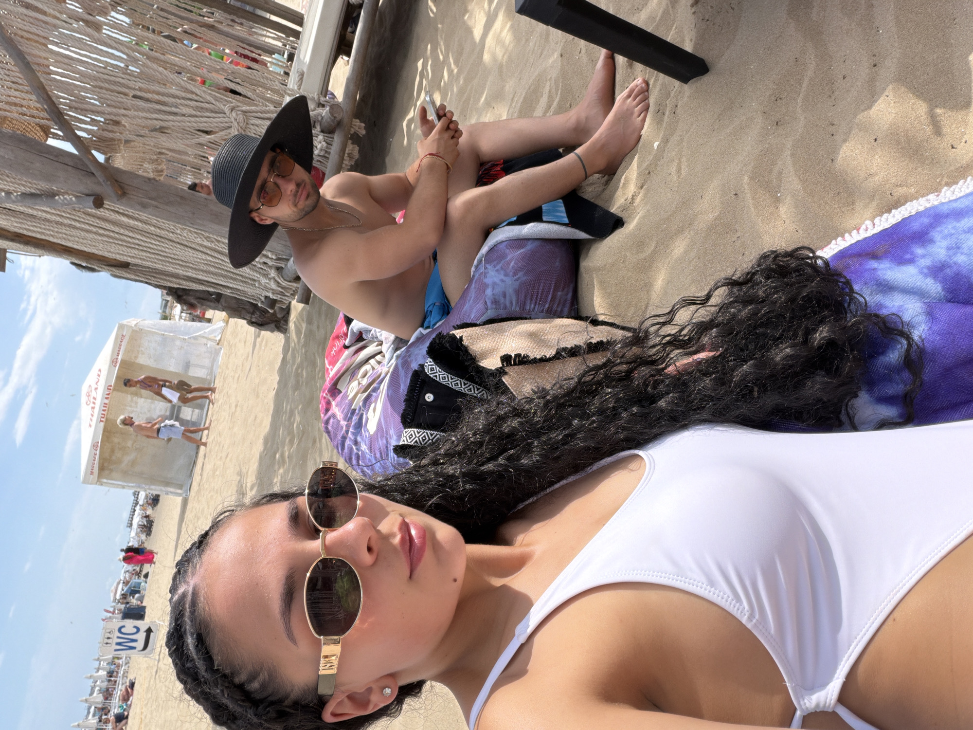 A medium shot captures two individuals on a sandy beach, likely within the city of Nesebar, Bulgaria, under a bright, sunny sky. In the foreground, a woman with braided dark hair and sunglasses wears a white swimsuit. To her right, and slightly further back, a man with sunglasses and a wide-brimmed hat sits on a towel, wearing blue swim trunks. He holds a smartphone in his right hand. Behind them, a white tent with "THAILAND" visible on it, along with beach umbrellas and loungers, indicates a resort or organized beach area. The sand is light brown, with some shadows cast by structures and people. The background shows a clear blue sky with scattered clouds.