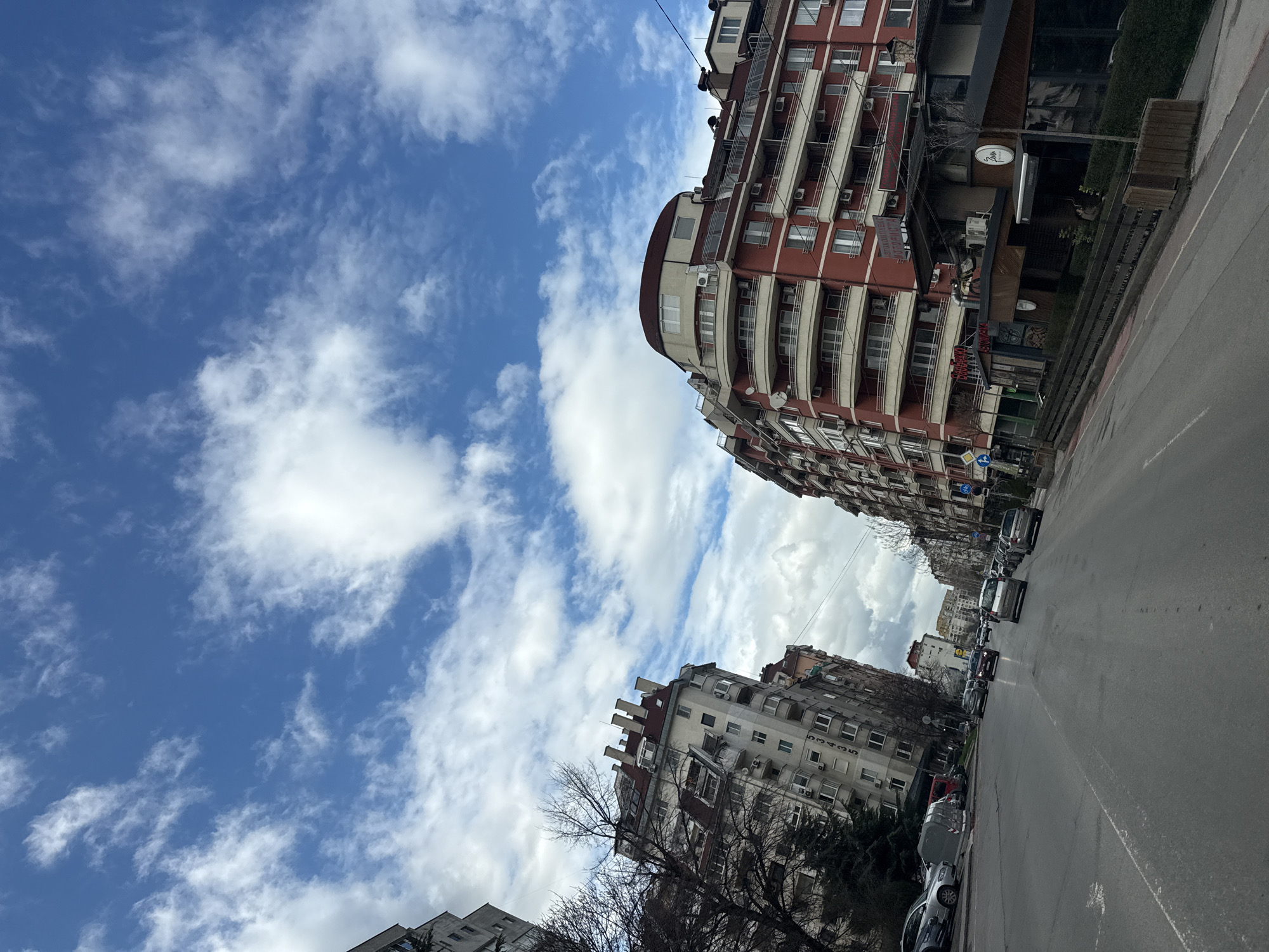 The image captures a street scene in Sofia, Bulgaria. The view is looking up the street, which is lined with buildings on both sides. The buildings are a mix of architectural styles, with some being taller and more modern, while others are older and more ornate. The sky is visible above the buildings and is blue with scattered white clouds. Cars are parked and moving on the street. The overall impression is of a typical urban environment in a European city.