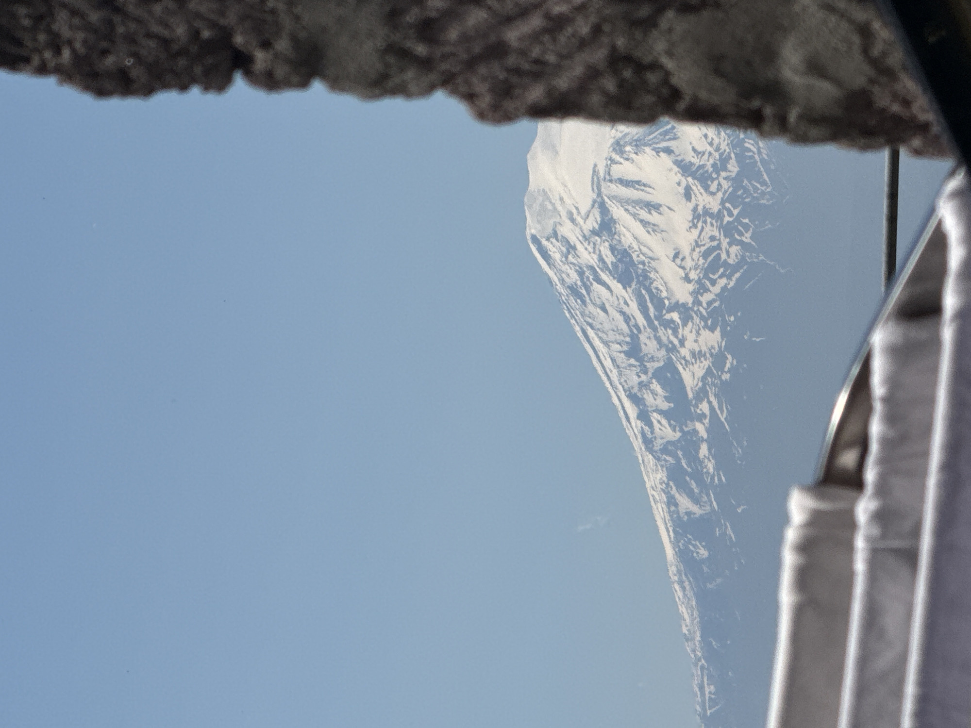 The image displays a serene outdoor scene, viewed through an opening or frame. The primary subject is a majestic, snow-capped mountain range, dominating the right half of the frame. The mountain features rugged terrain with prominent peaks and valleys, covered extensively in white snow contrasted with darker rock faces. A clear, light blue sky fills the left side and upper portion of the frame, indicating a bright daytime setting with good weather.
In the immediate foreground, a dark, textured, and rough-edged object partially obscures the top of the view, resembling an overhang, a roof, or a rocky ledge. Along the bottom and right edges, a blurry, light-colored object with vertical folds and a metallic, curved element is visible, suggesting a window frame, a curtain, or an interior part of a vehicle or structure from which the photograph was taken.
There are no people or discernible activities taking place other than the act of observing the mountain view. The time of day appears to be daytime due to the bright blue sky. There is no visible text in the image.