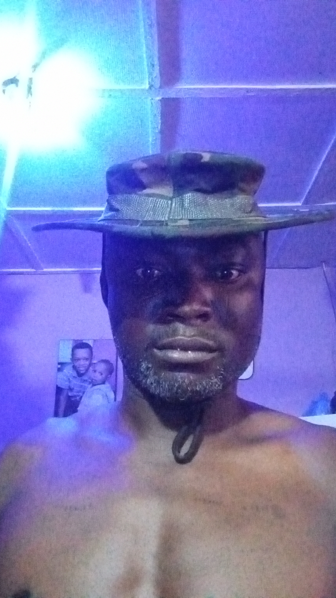 A shirtless man with a salt-and-pepper beard is looking directly at the camera. He is wearing a camouflage hat with a wide brim. Behind him is a photograph of two young children and the ceiling has a grid-like pattern. The location is not discernible from the image alone. There is no basis to determine if it is in the city of Sabon Tasha, Nigeria. The lighting is a blue-purple hue.