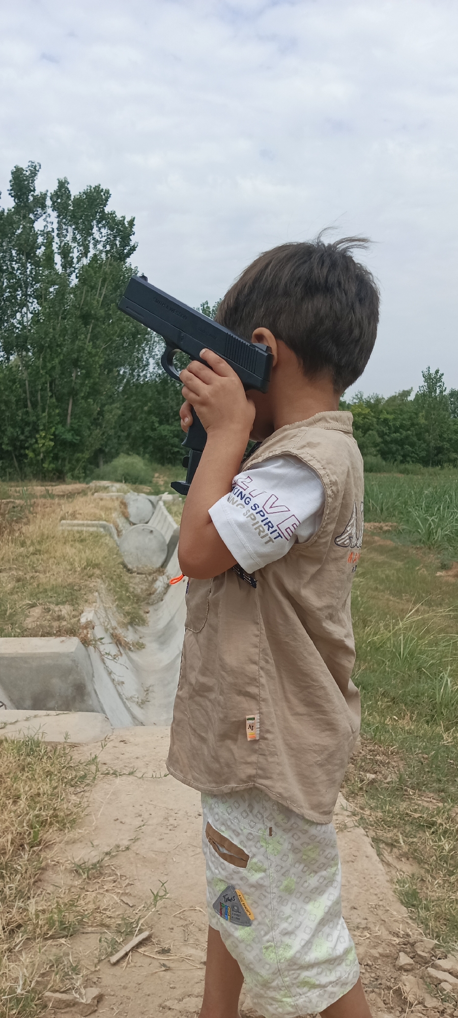 A boy, seen from the side and from the back, stands outdoors holding a black toy gun to his right ear and temple. He wears a tan vest over a white t-shirt with text and a pair of patterned shorts. The background consists of trees, dry grass, and a concrete structure, possibly a drainage ditch, indicative of a rural or semi-urban setting within Katlang Tehsil, Pakistan. The sky is overcast.